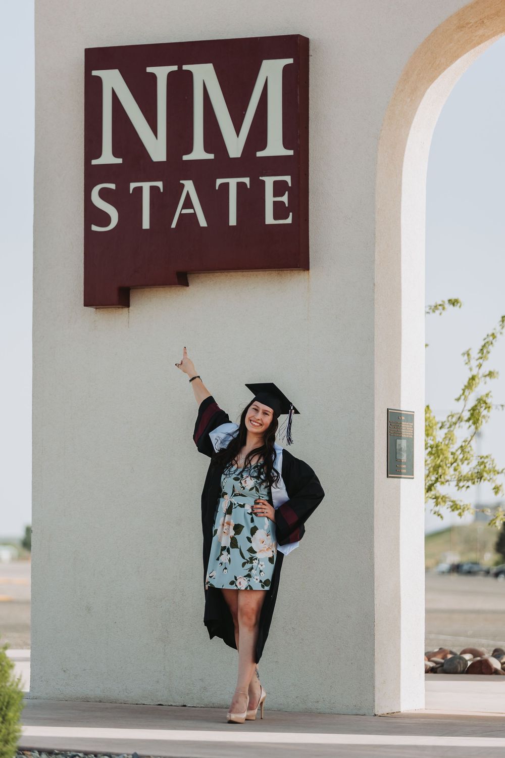 Graduate celebrating in graduation cap and gown with decorative floral dress under concrete archway at New Mexico State.
