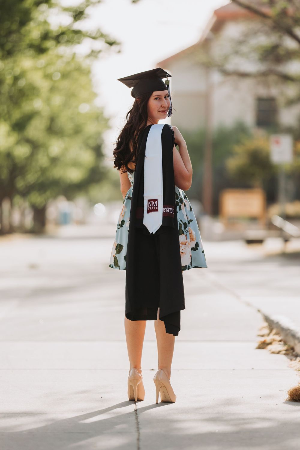 Graduate in cap and gown walking down sunny sidewalk wearing black dress and high heels with floral tote bag.