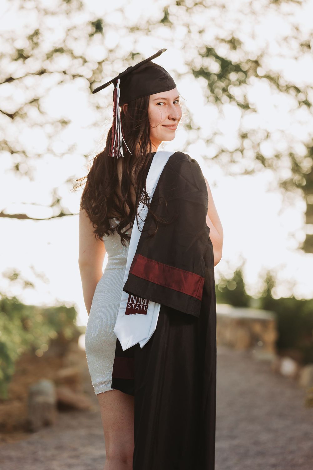 A graduate wearing a cap and gown poses outdoors during golden hour with a warm bokeh background.