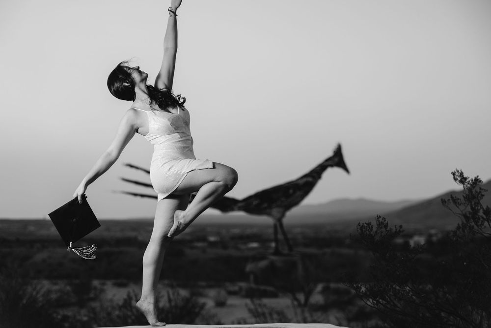 A graceful dancer in a white dress leaps joyfully against a mountainous landscape in black and white.