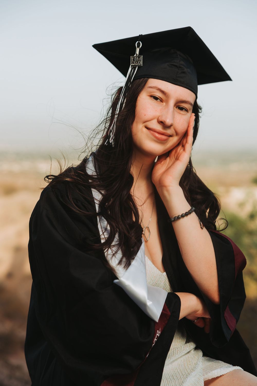 Graduate in black cap and gown poses outdoors against mountain backdrop during sunset photo session.