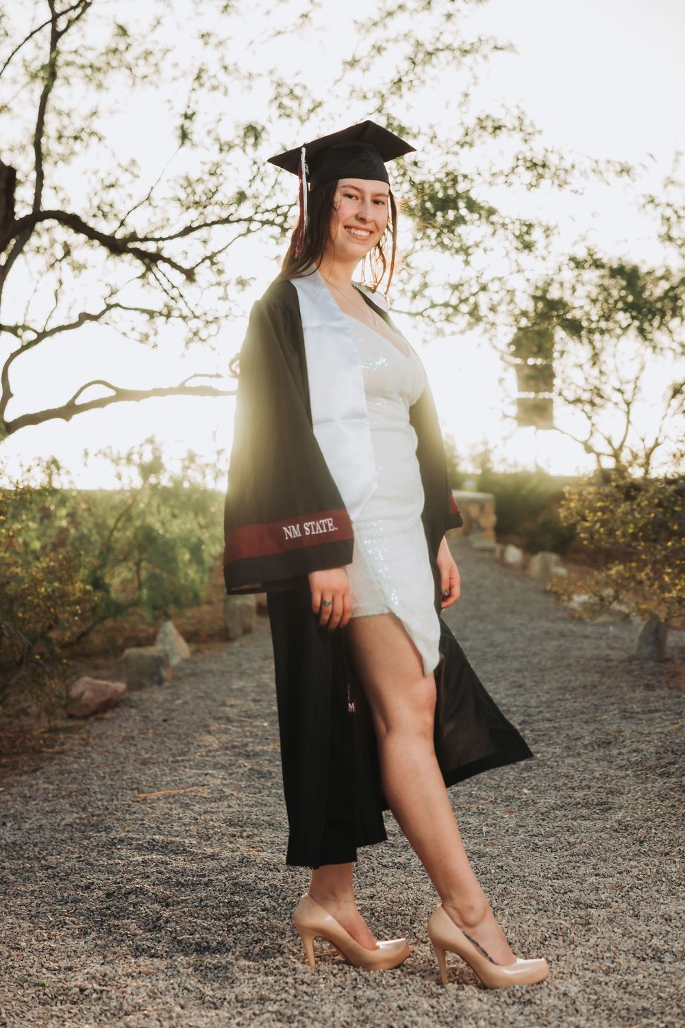 A graduate in a white dress and black cap and gown poses on a gravel path at sunset.