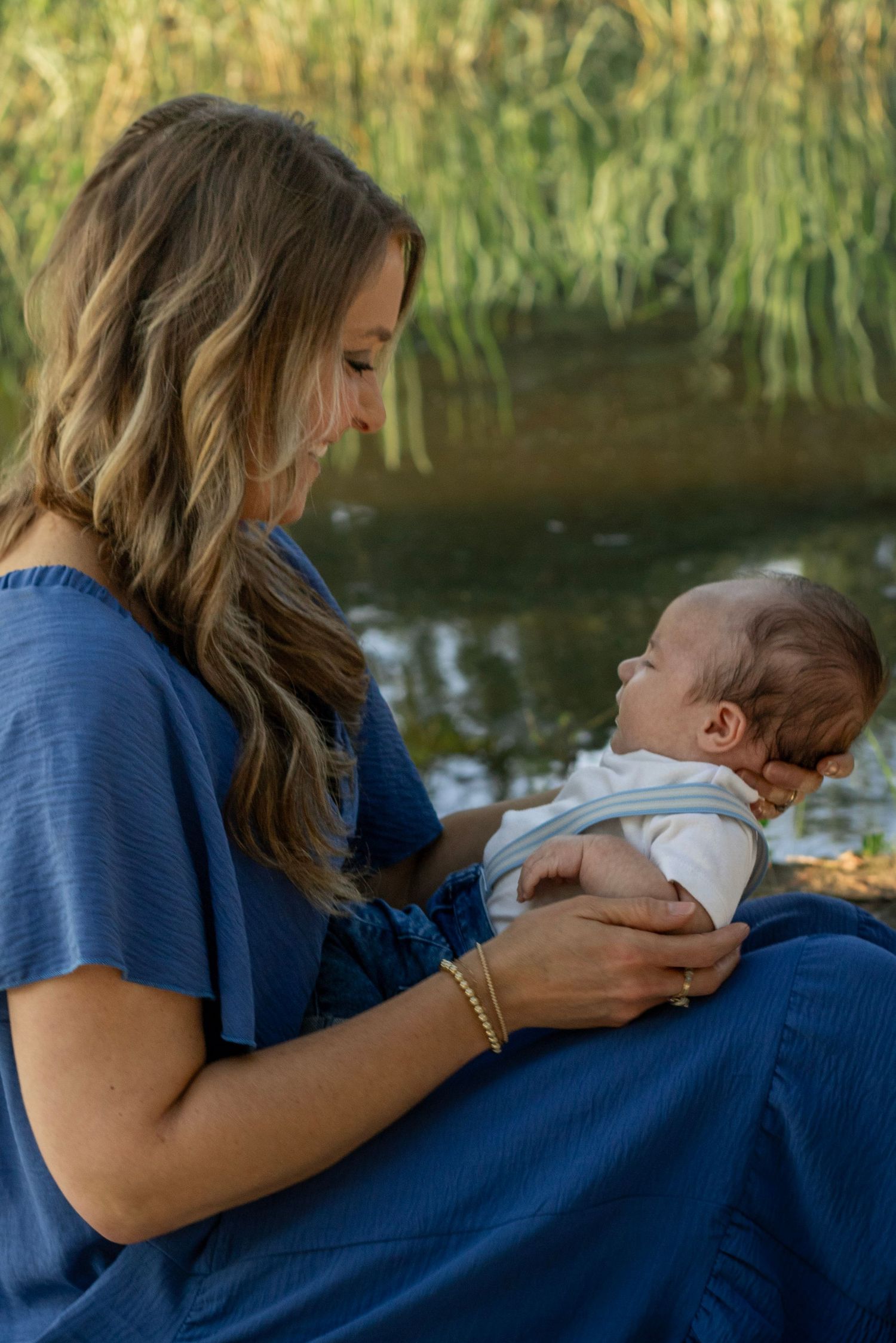 Mother sitting near a Lake Wateree with newborn in blue dress, willow trees reflecting in the water backdrop.
