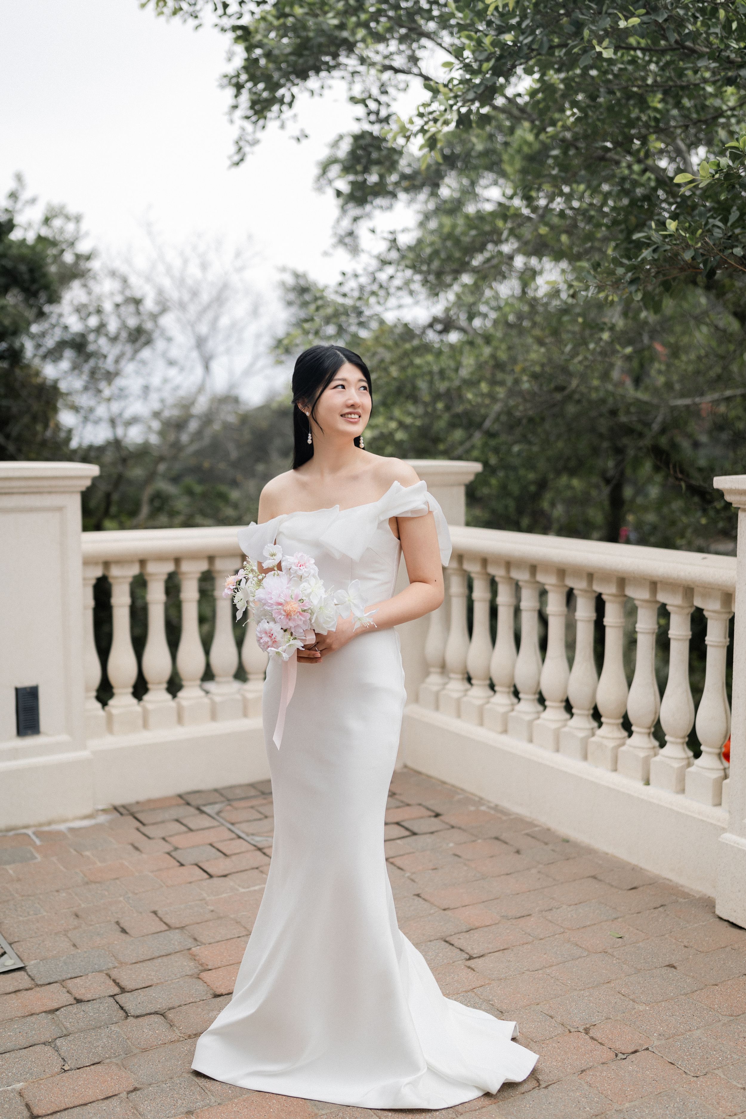 Smiling bride in off-shoulder white fitted gown holding pastel bouquet on a terrace with balustrade.