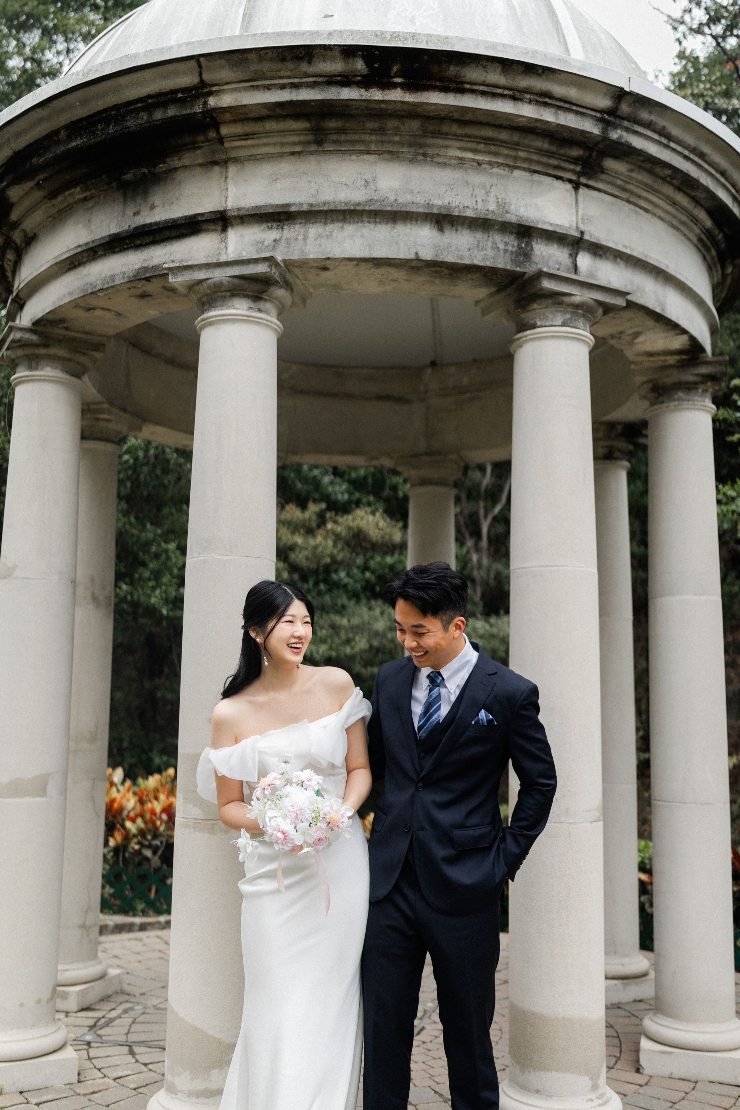 Smiling bride and groom pose together under a grand white columned rotunda surrounded by greenery.