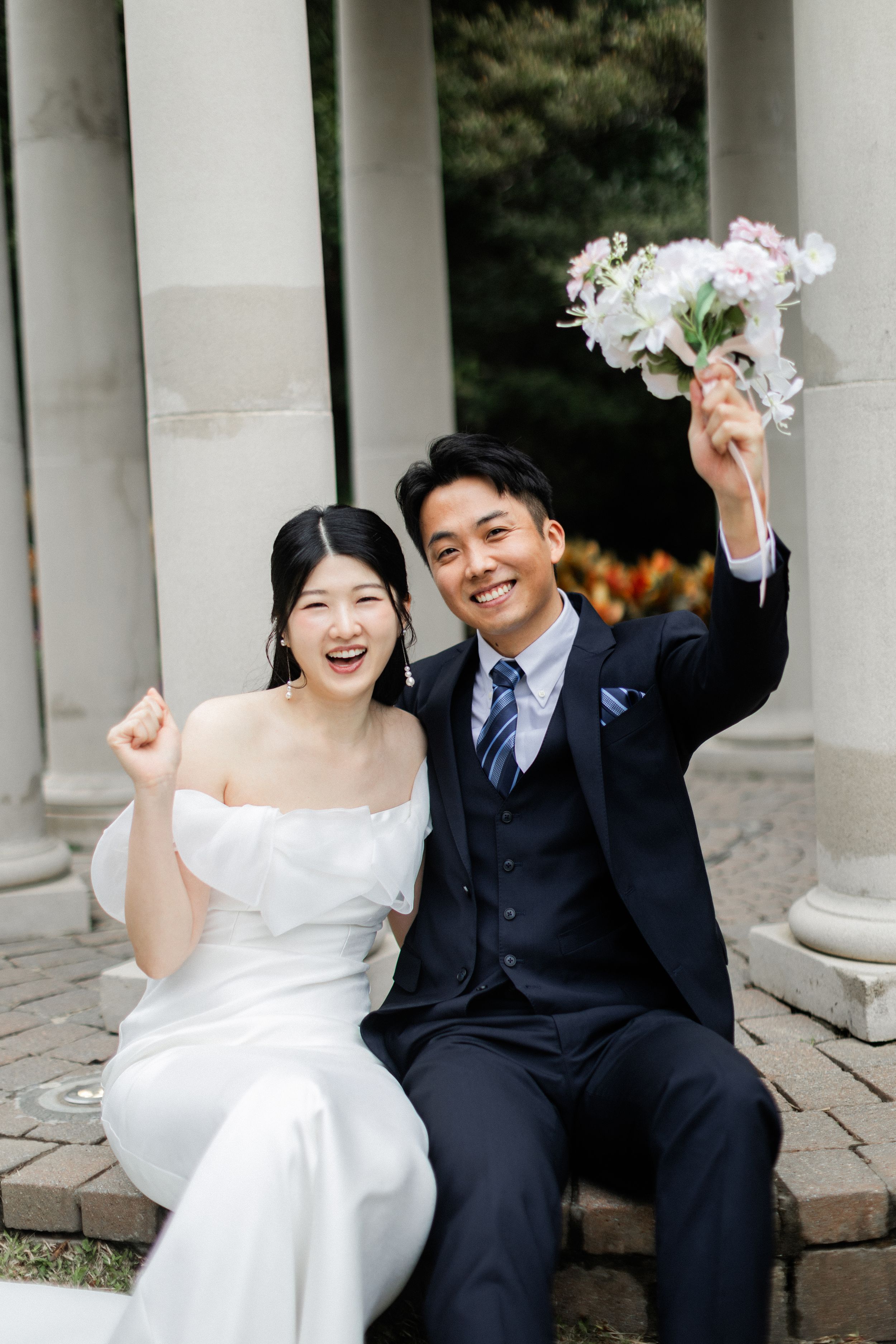 Joyful bride and groom celebrate with raised fists and bouquet while seated at a classical stone structure.