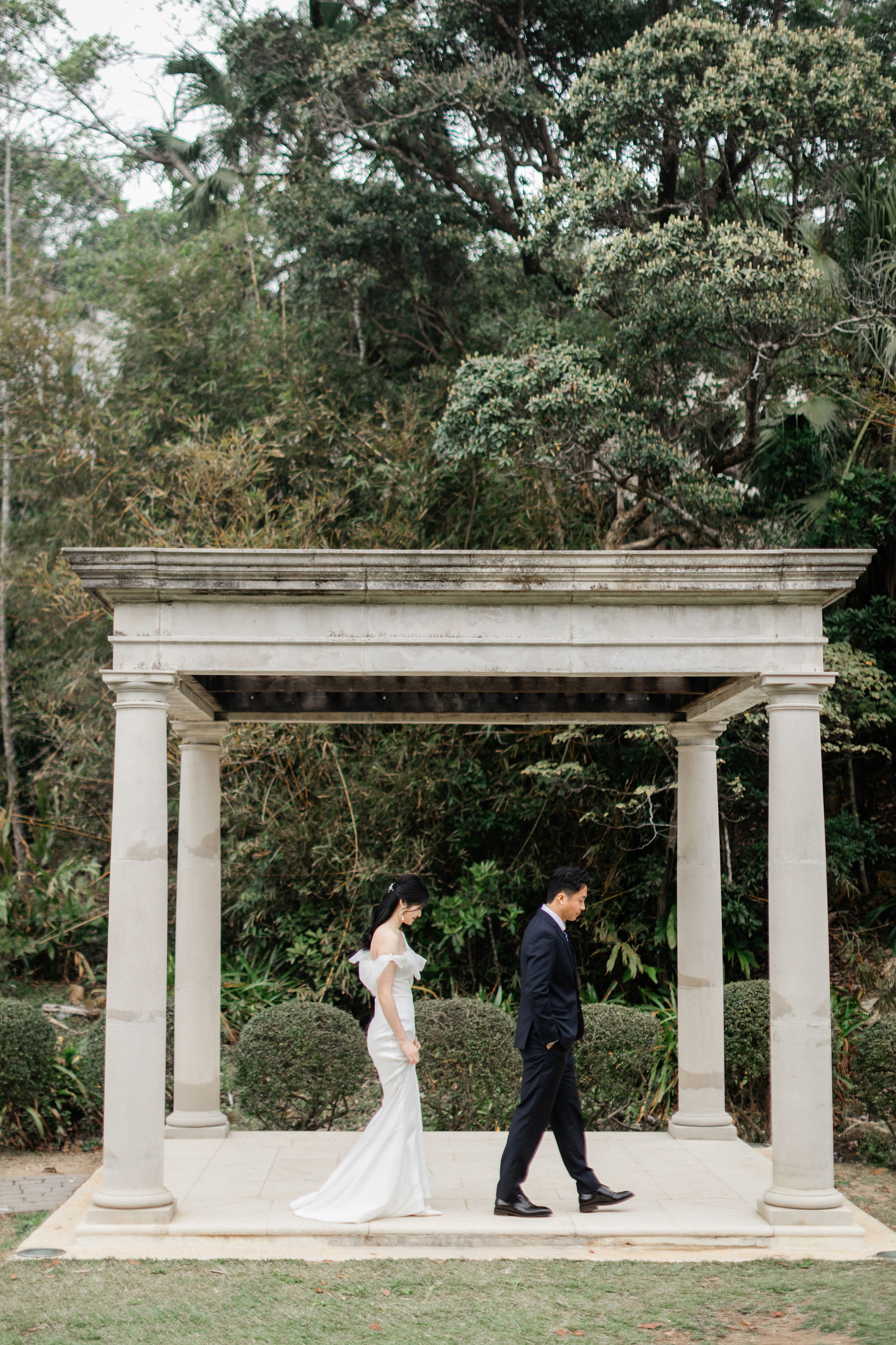 Elegant bride and groom walk beneath a neoclassical stone archway with columns in a garden setting.