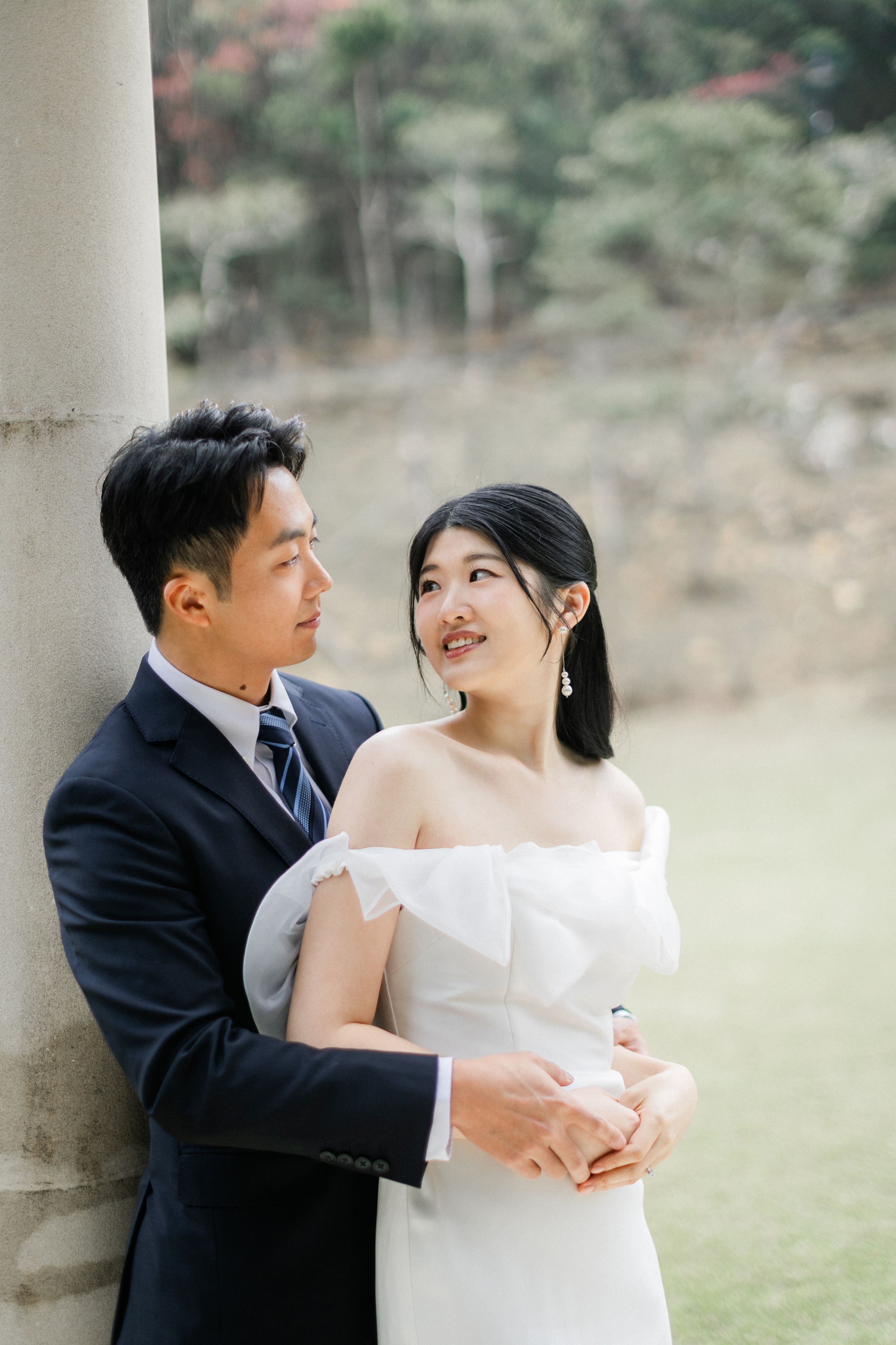 Groom tenderly embraces bride from behind as they share a loving gaze beside a stone column outdoors.