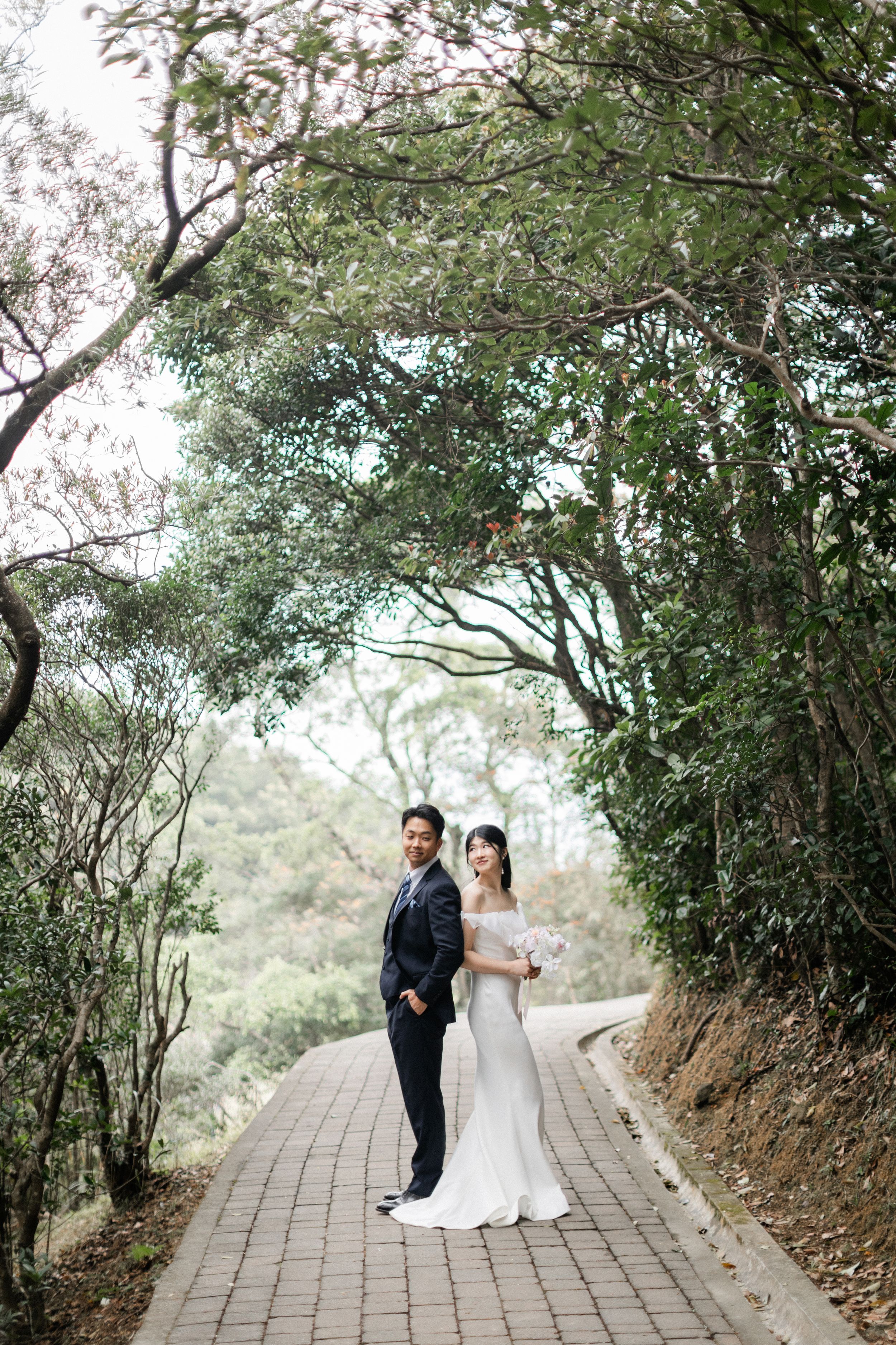 Bride and groom stand together on a tree-lined brick pathway surrounded by lush green forest canopy.