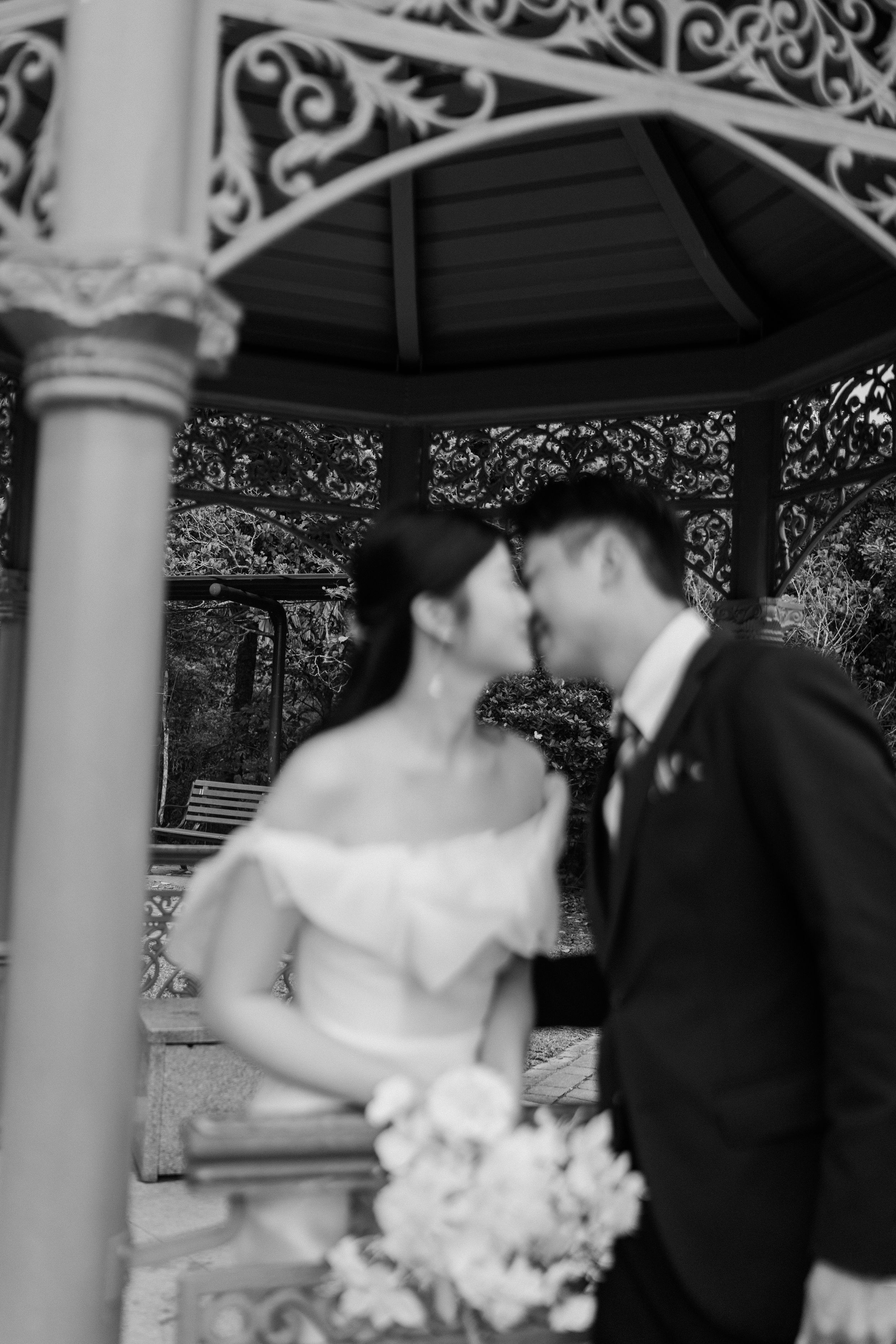 Black and white romantic photo of bride and groom sharing a kiss inside an ornate wrought iron gazebo.