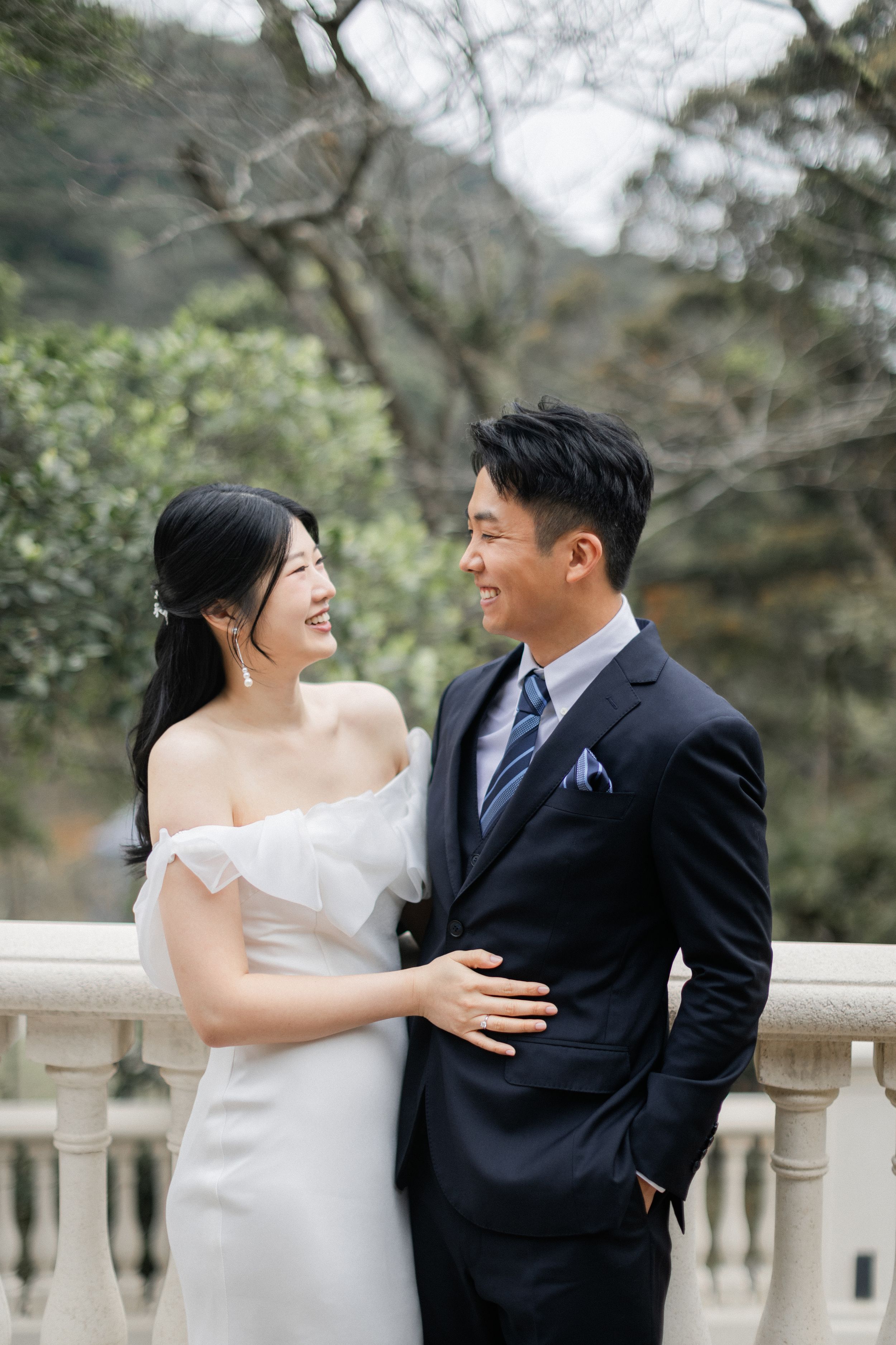 Laughing couple in wedding attire embracing on a terrace, groom in navy suit, bride in white gown.