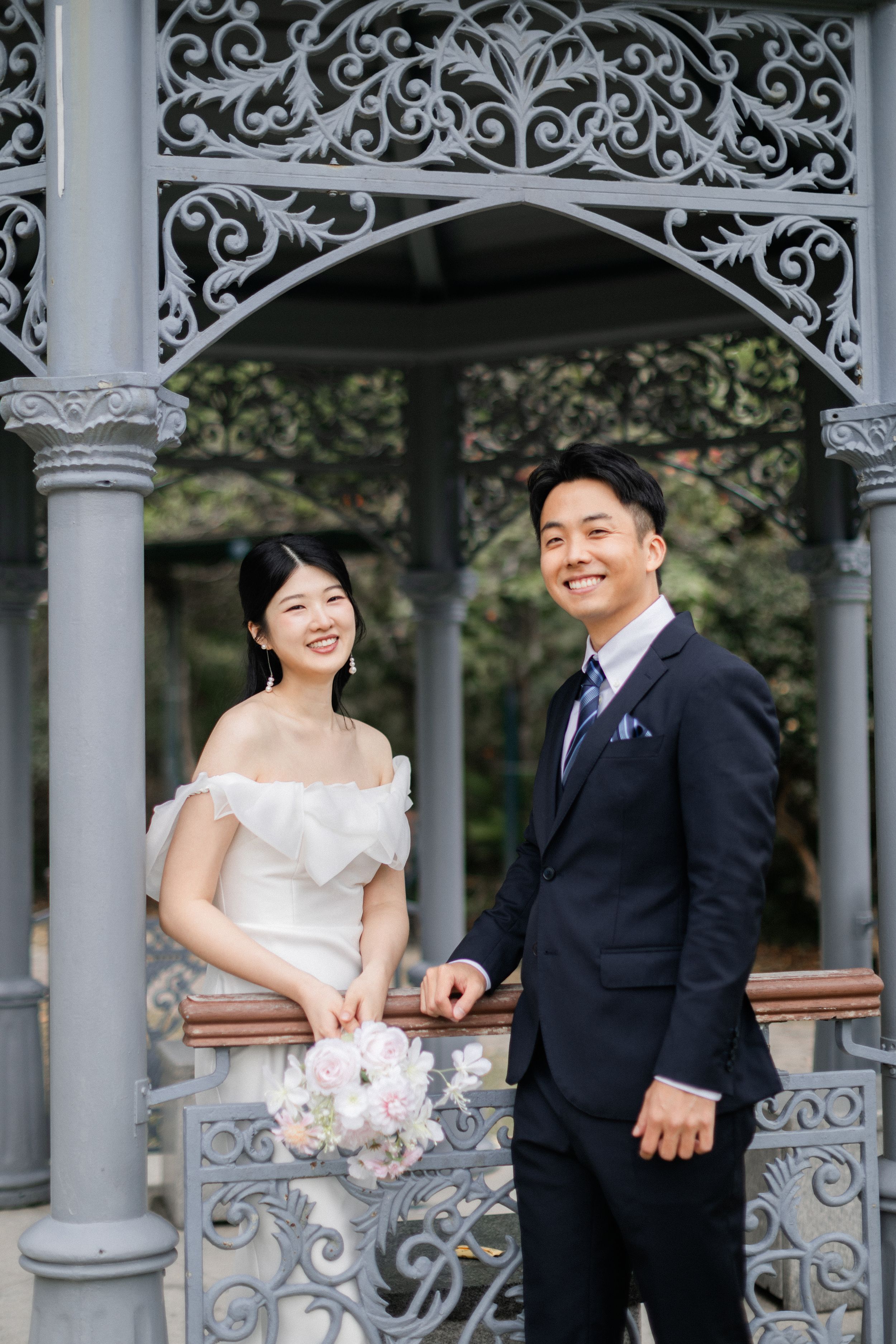 Smiling bride and groom pose inside a decorative grey wrought iron gazebo holding a pastel bouquet.