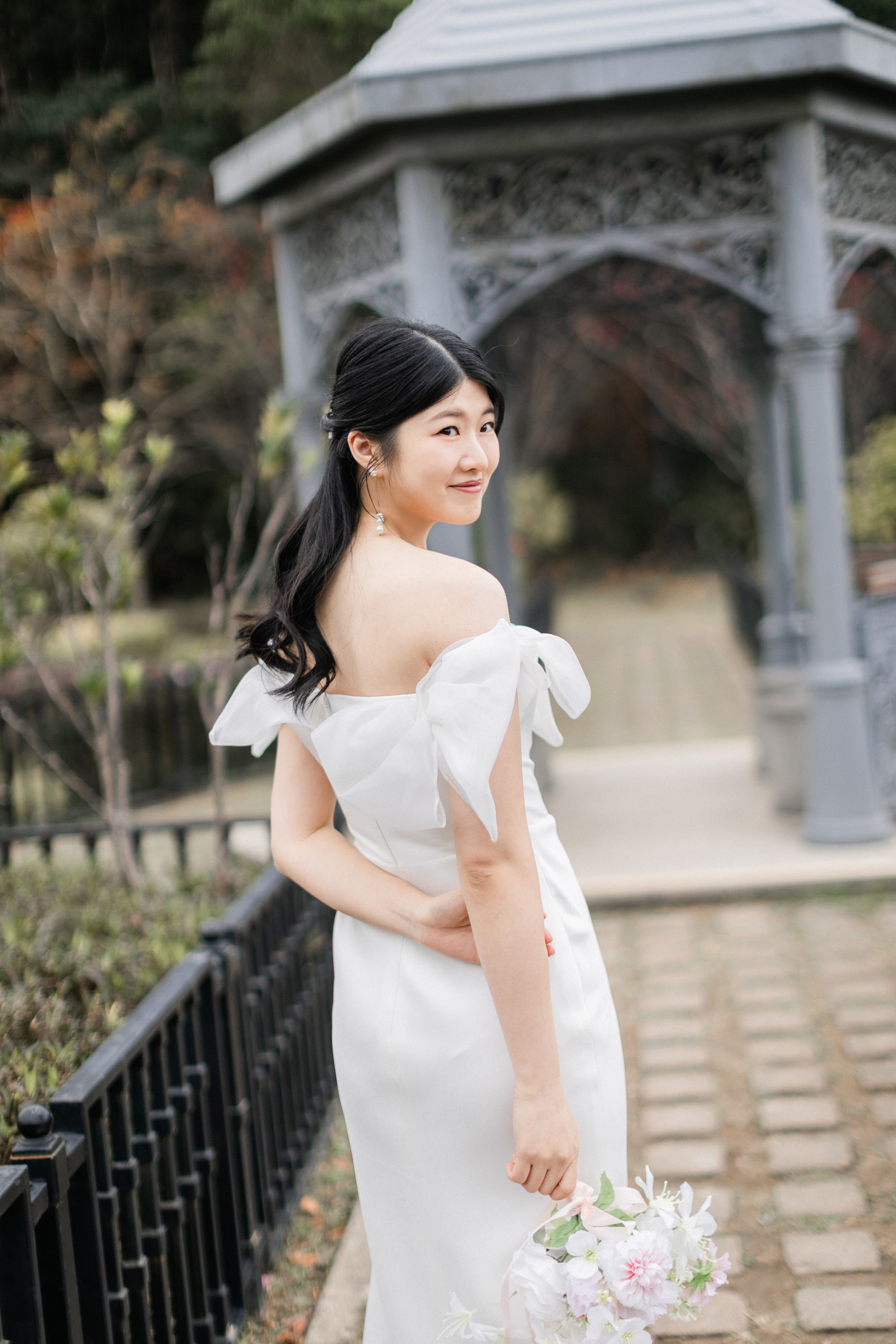 Elegant bride in off-shoulder white gown glances back gracefully with a grey ornate gazebo in background.