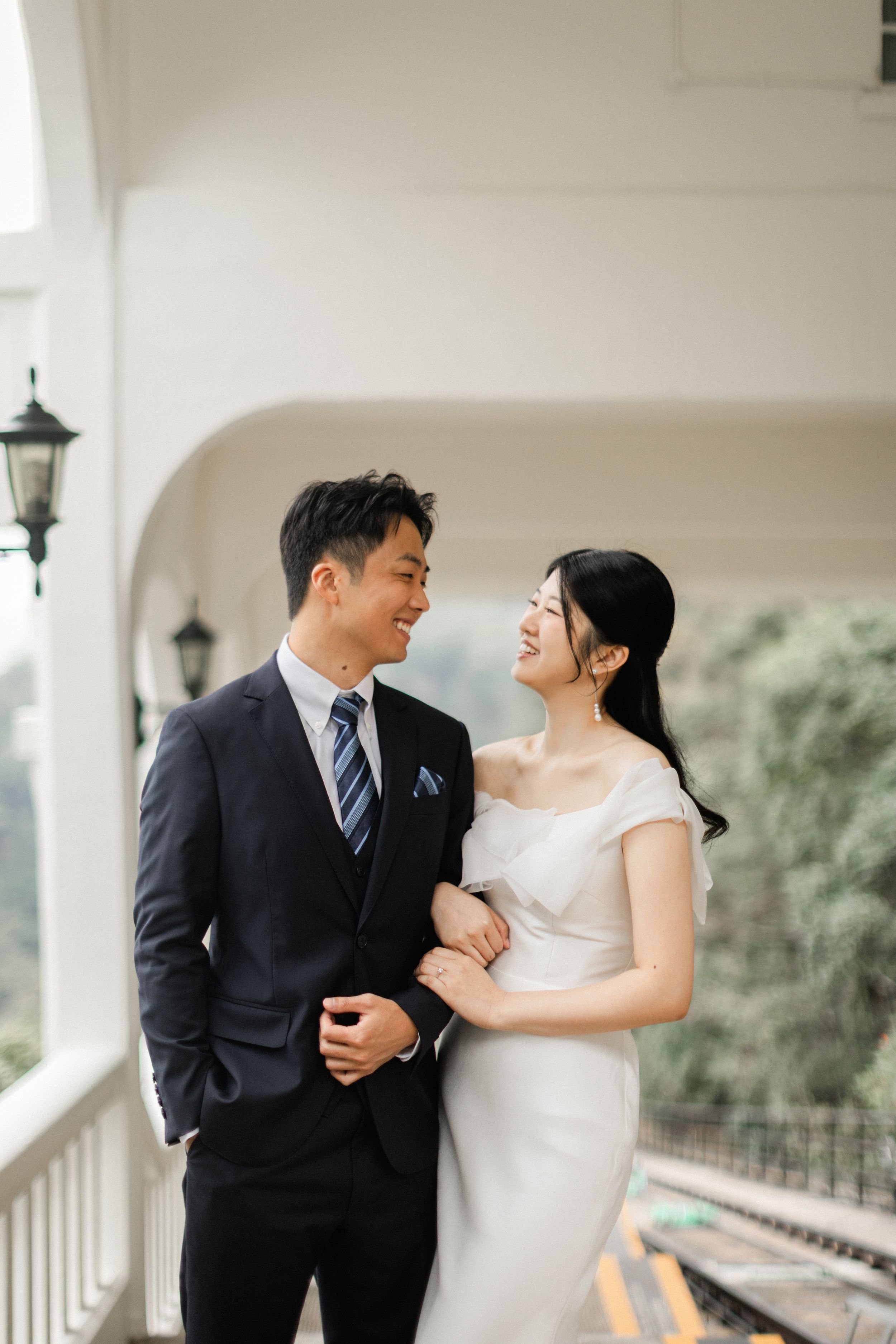 Smiling Asian couple in wedding attire share a romantic moment on a white colonial balcony.