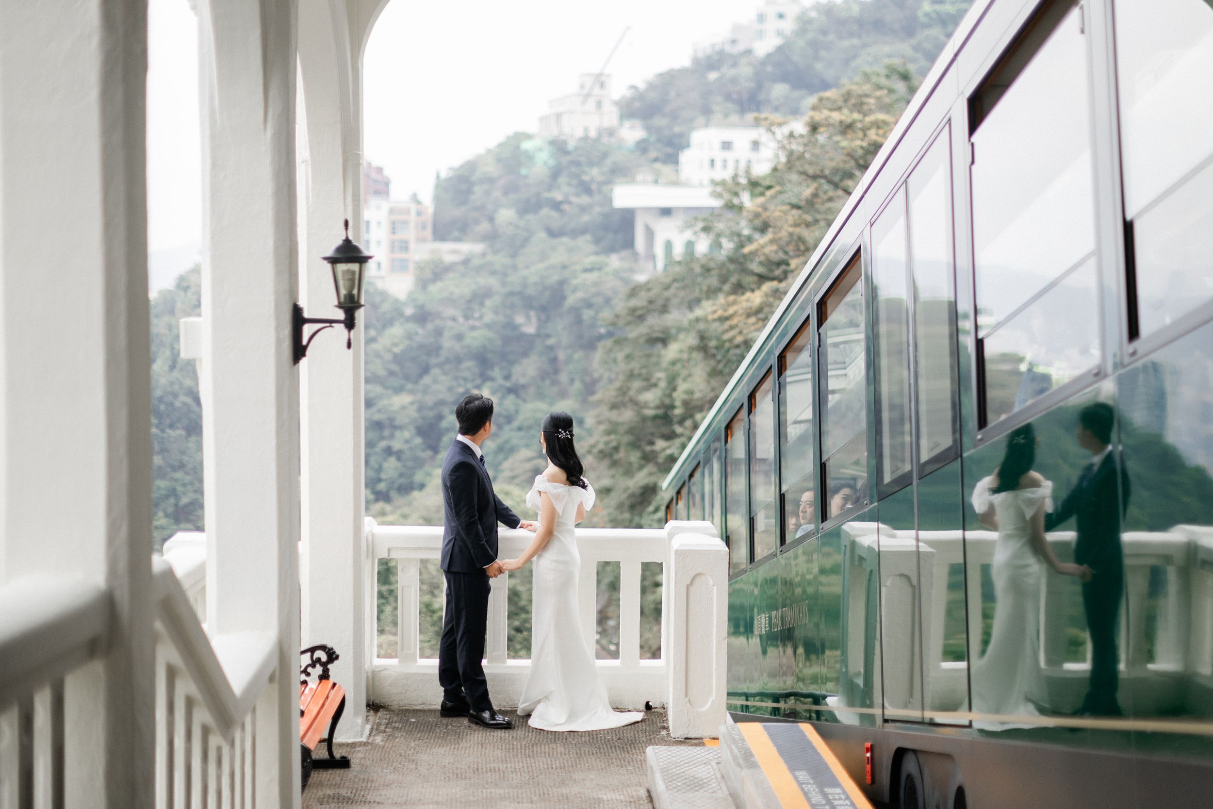 Wedding couple stands on elegant white balcony beside a green tram in Hong Kong.