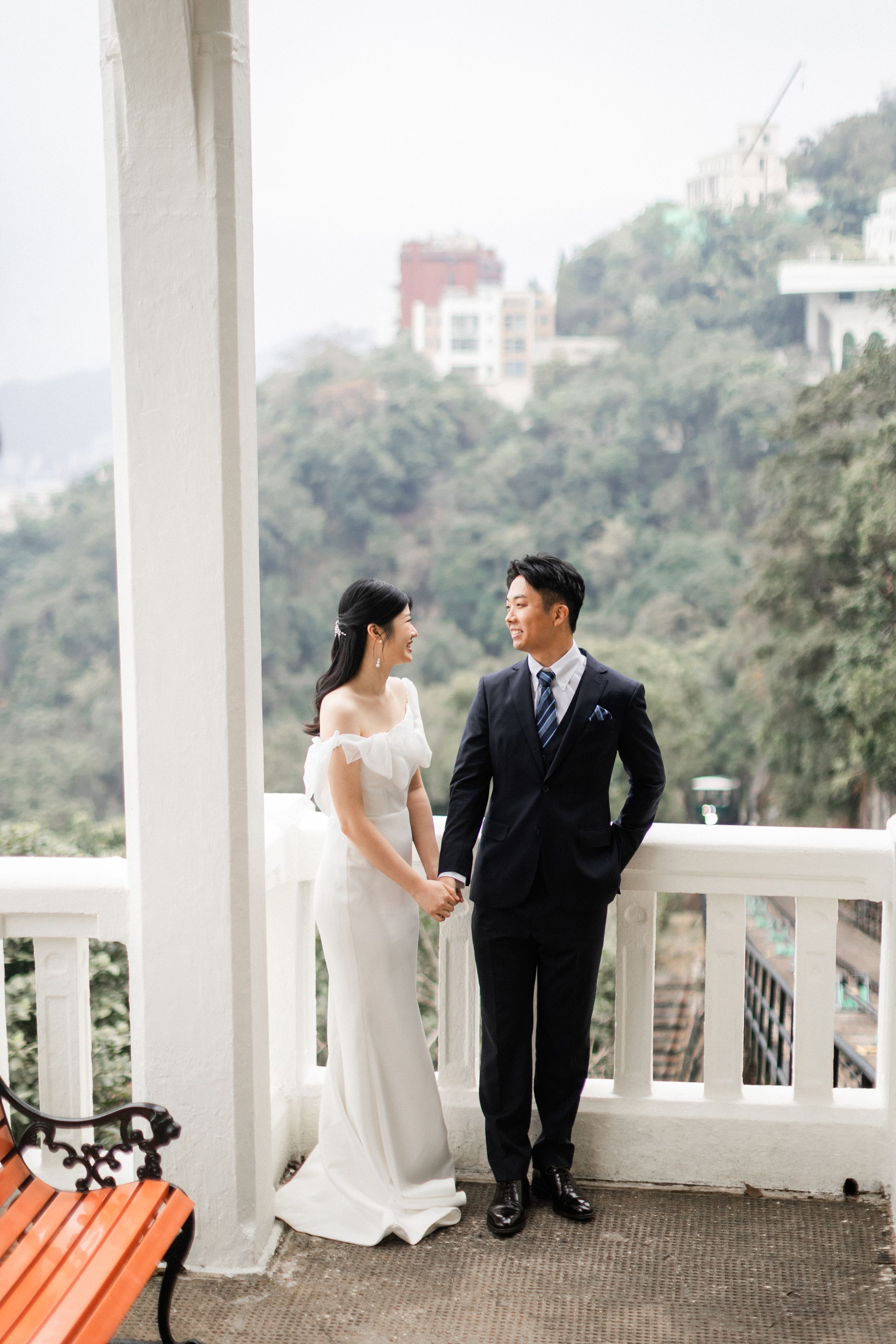 Asian bride and groom hold hands on a white colonial veranda with green hills behind.