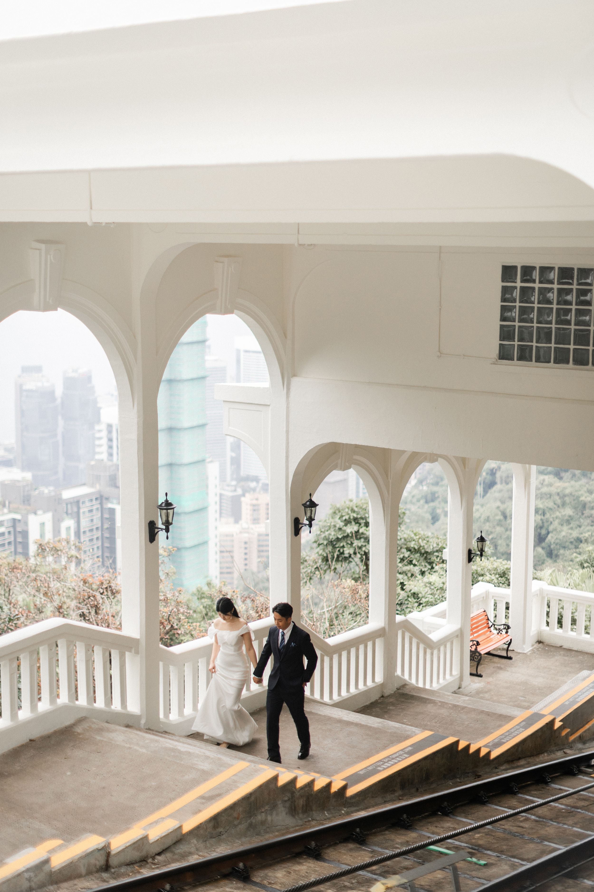 Wedding couple walks hand in hand through white arched colonial building with city views.
