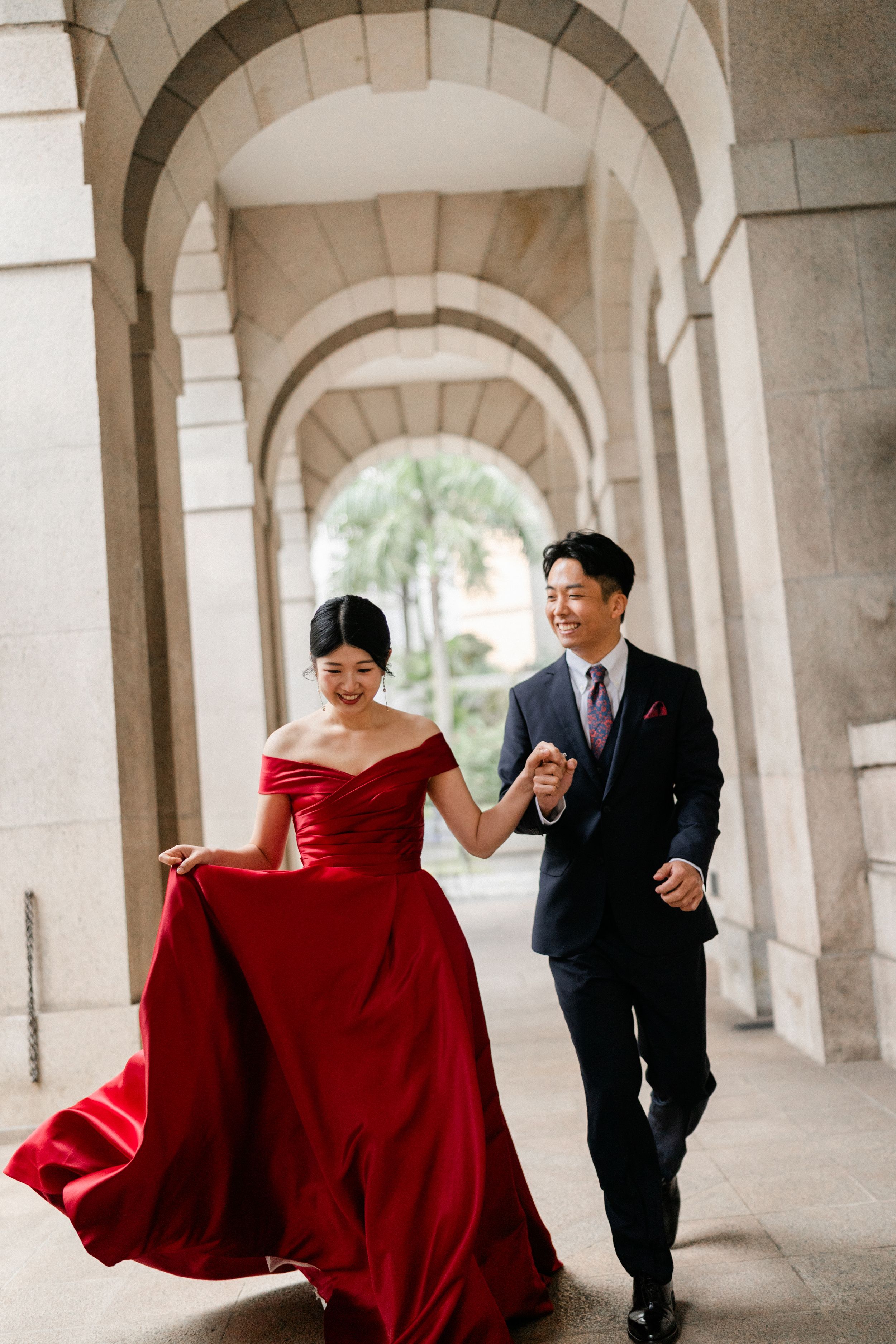 Joyful couple runs through stone archway colonnade, bride in flowing red off-shoulder gown.