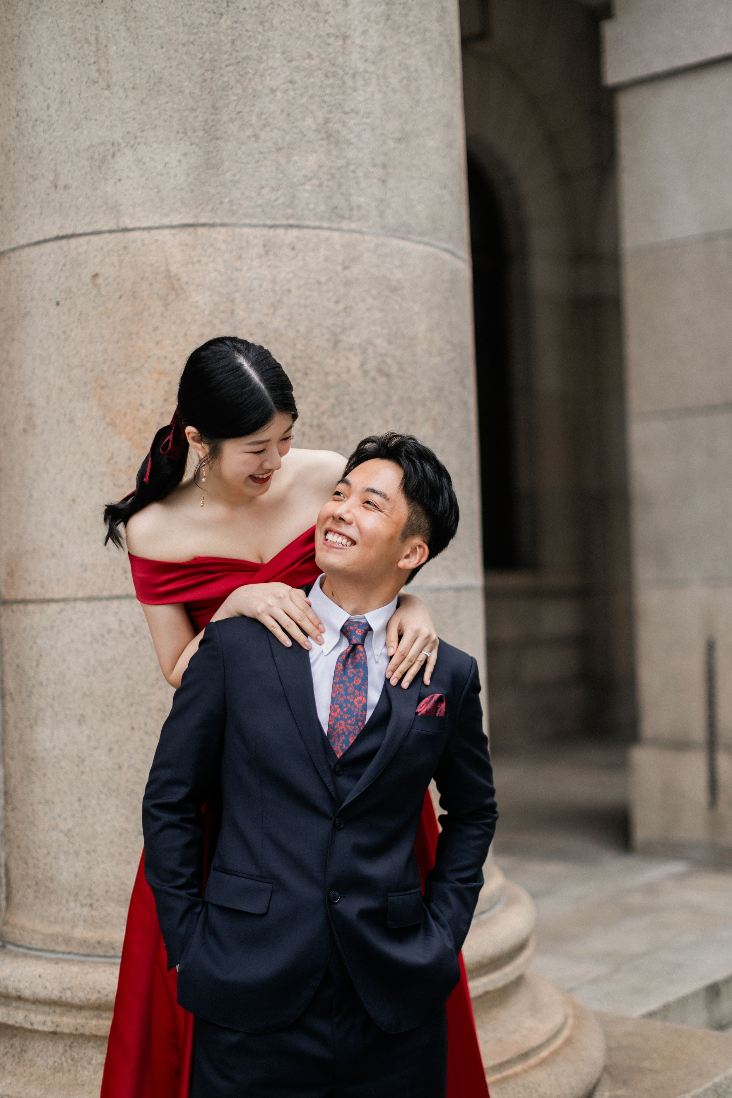 Laughing bride in red gown embraces groom from behind beside a large stone column.