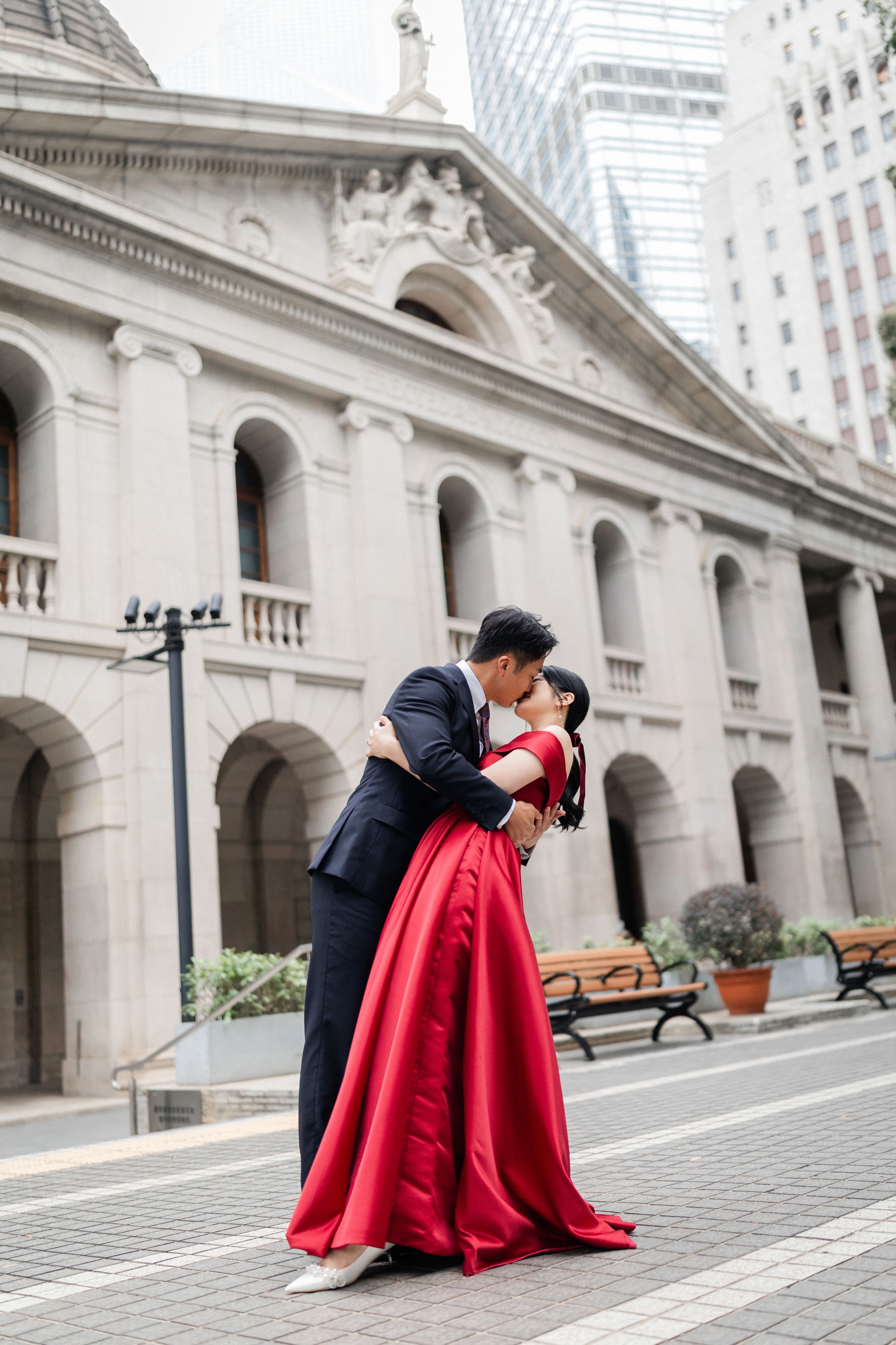Groom dips bride in red ballgown for a kiss outside a grand neoclassical building.
