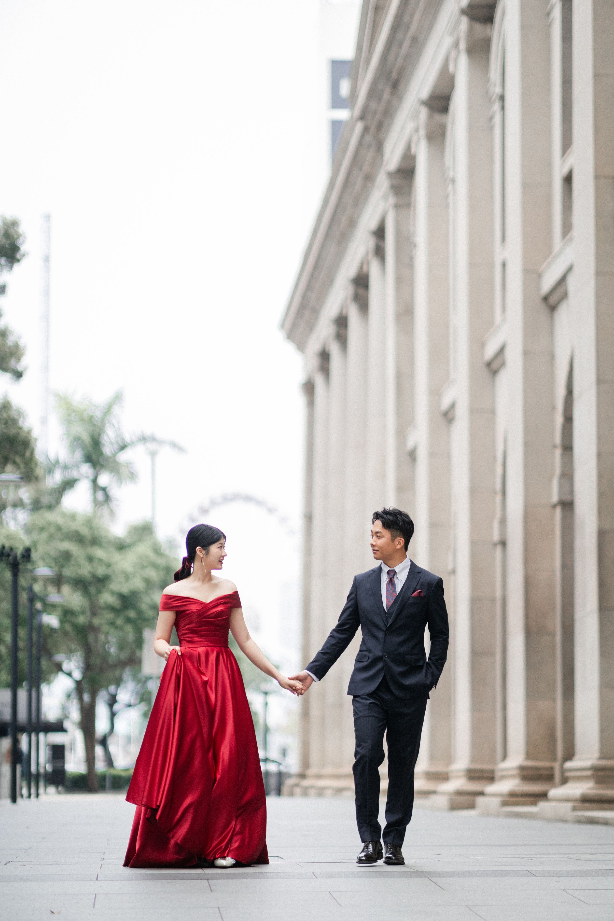 Couple in red gown and dark suit holding hands walking past classical stone building columns.