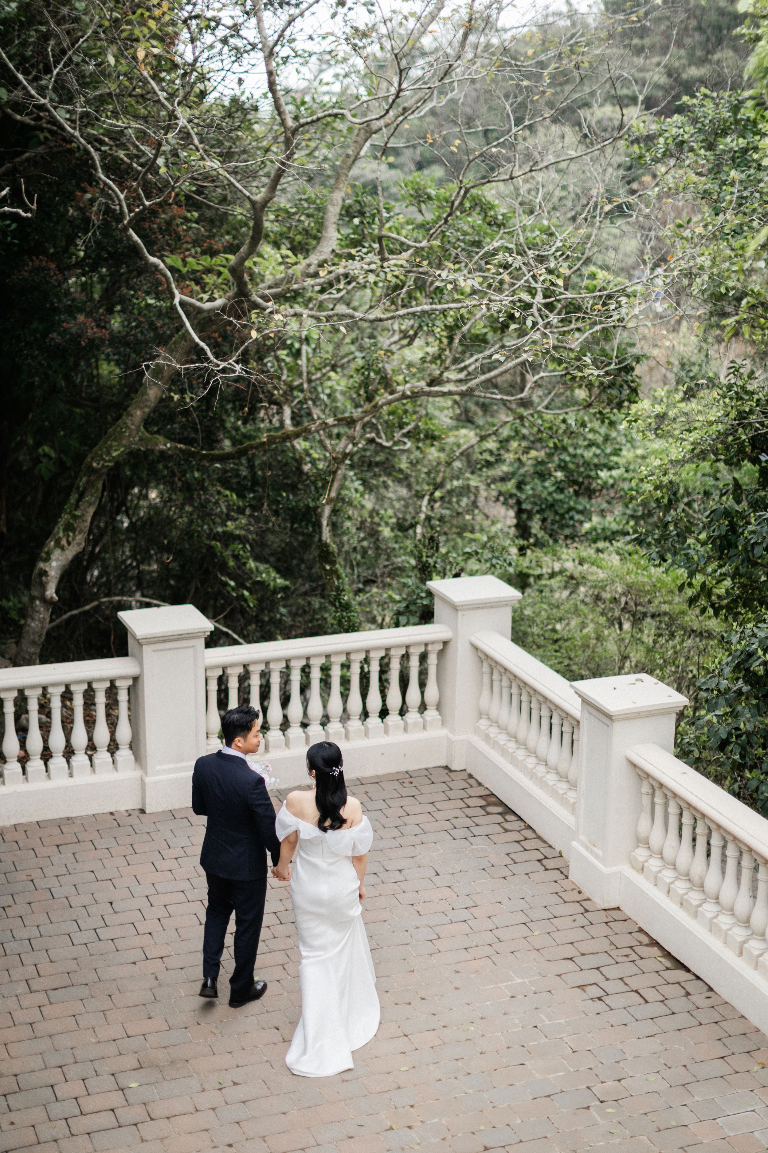 Bride and groom standing hand in hand on a brick terrace overlooking a lush green garden.