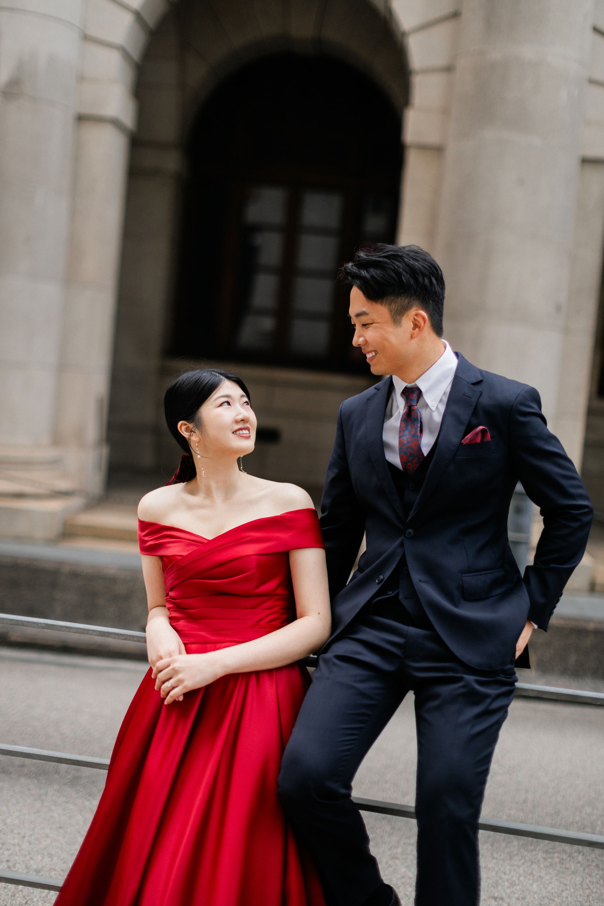 Smiling couple in red off-shoulder gown and navy suit sharing a joyful moment on stone steps.