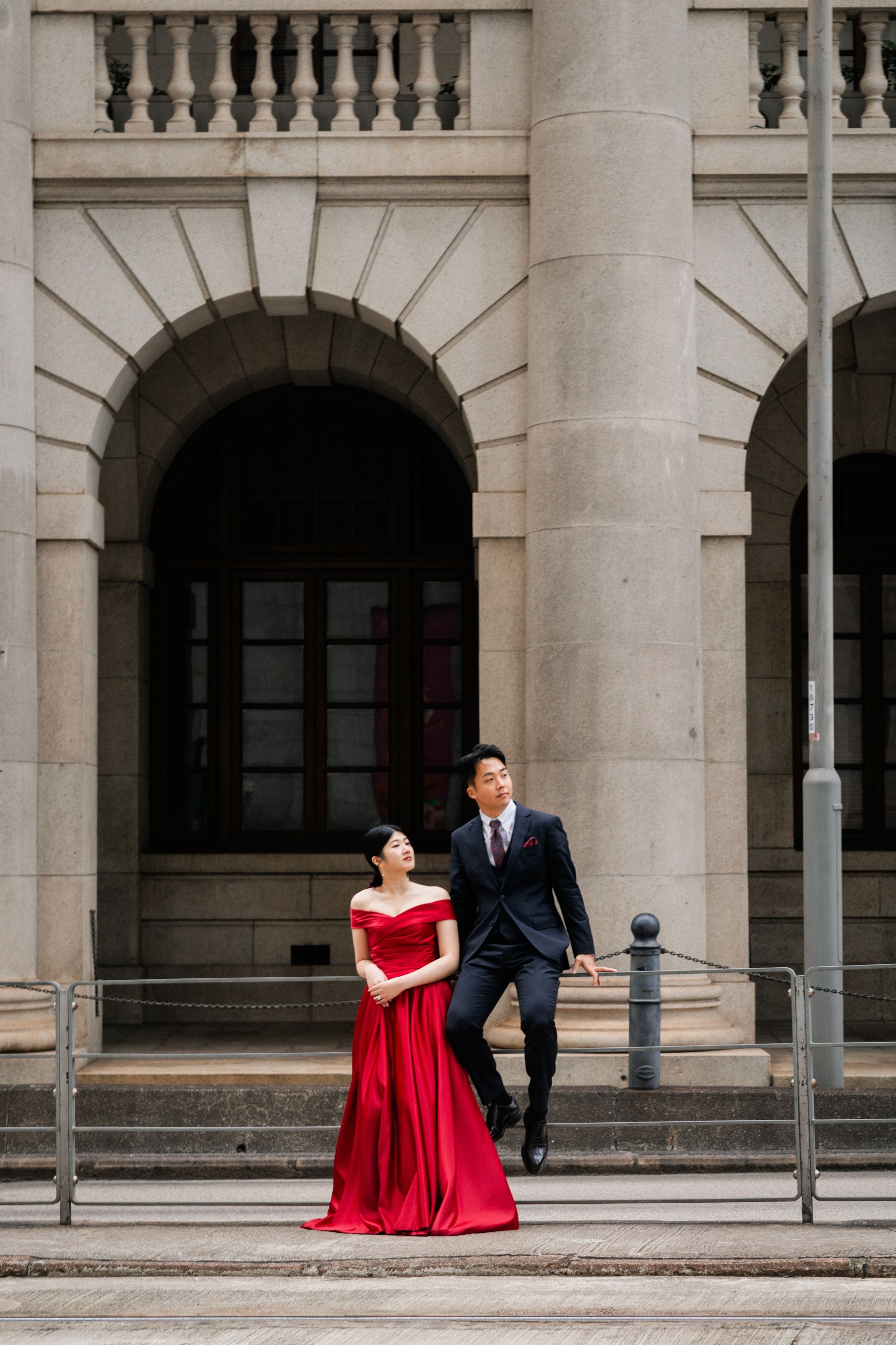 Couple in red ball gown and dark suit posing by grand arched stone building entrance.