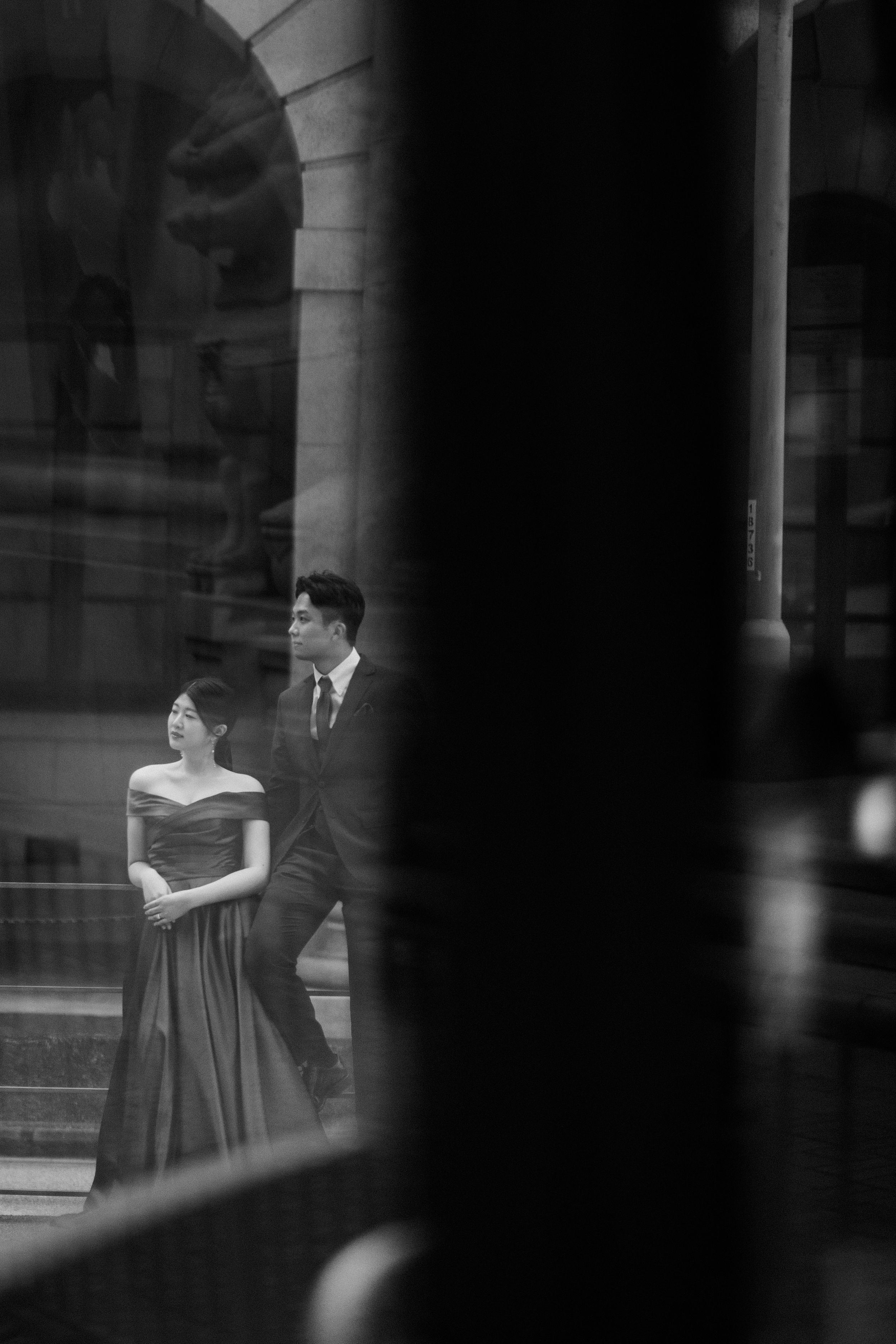 Black and white photo of elegant couple in formal wear walking past ornate building archway.