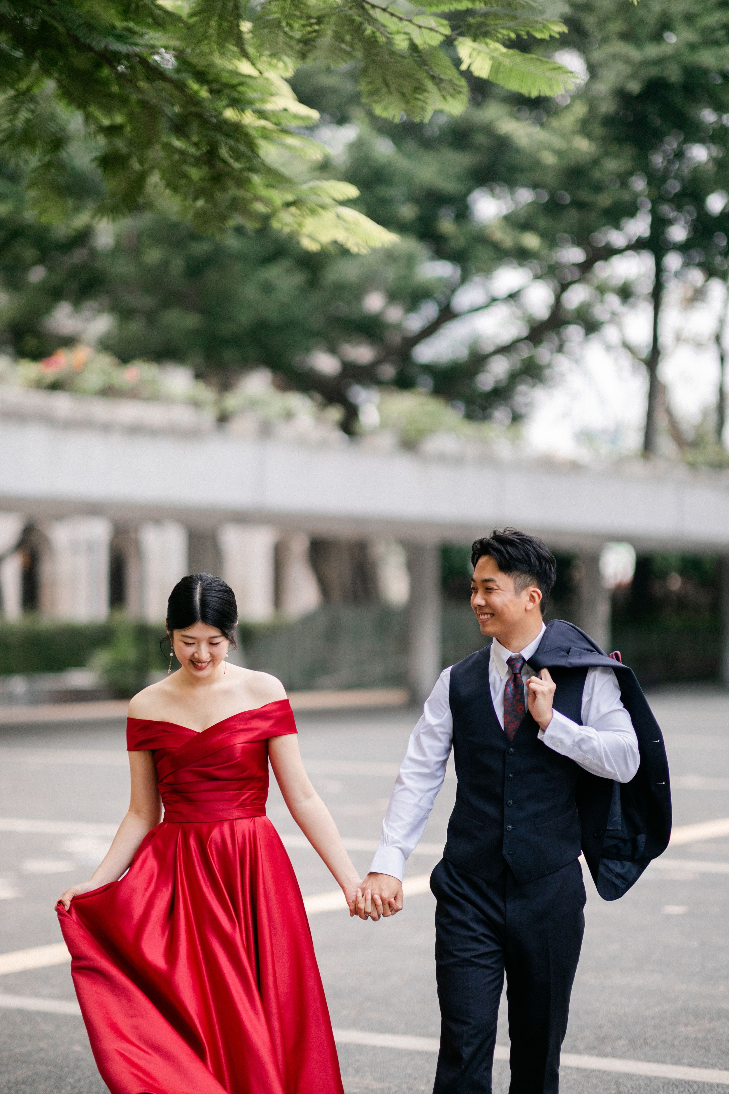Couple holding hands walking outdoors, woman in flowing red satin gown, man carrying suit jacket.