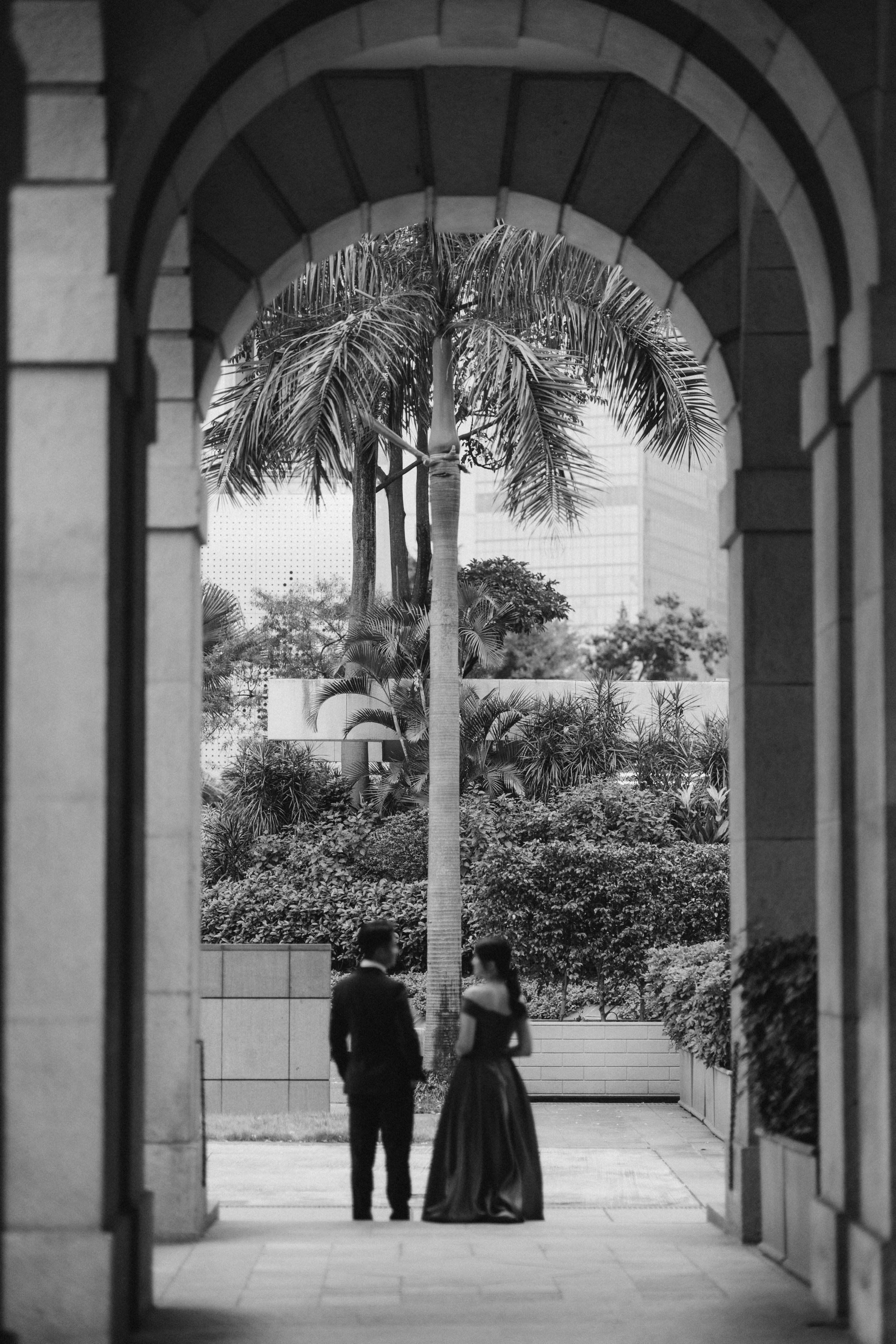 Black and white photo of couple standing under stone archway with tropical palm tree backdrop.