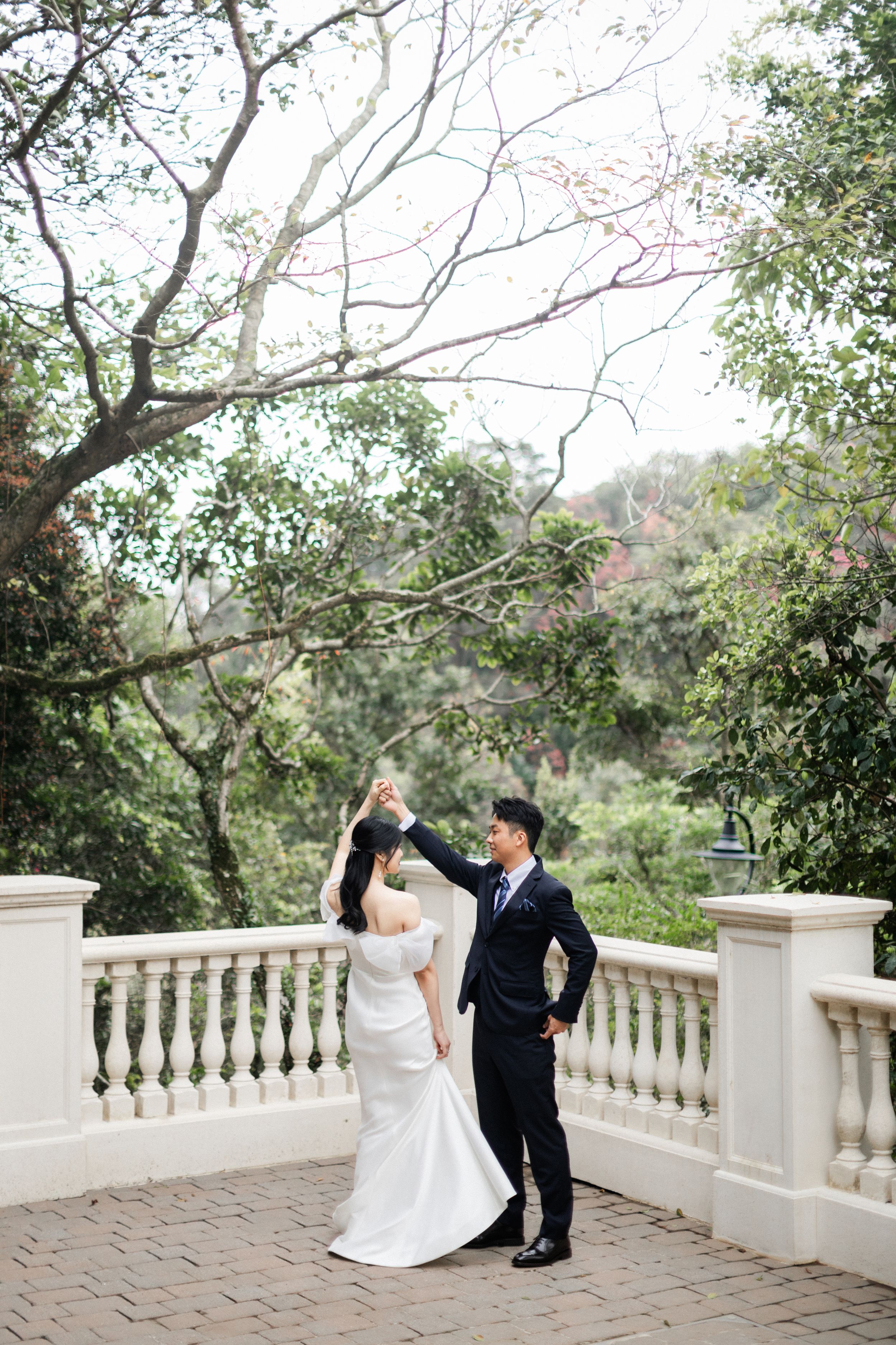 Groom spinning bride in a joyful dance on a terrace with trees and white balustrade in background.