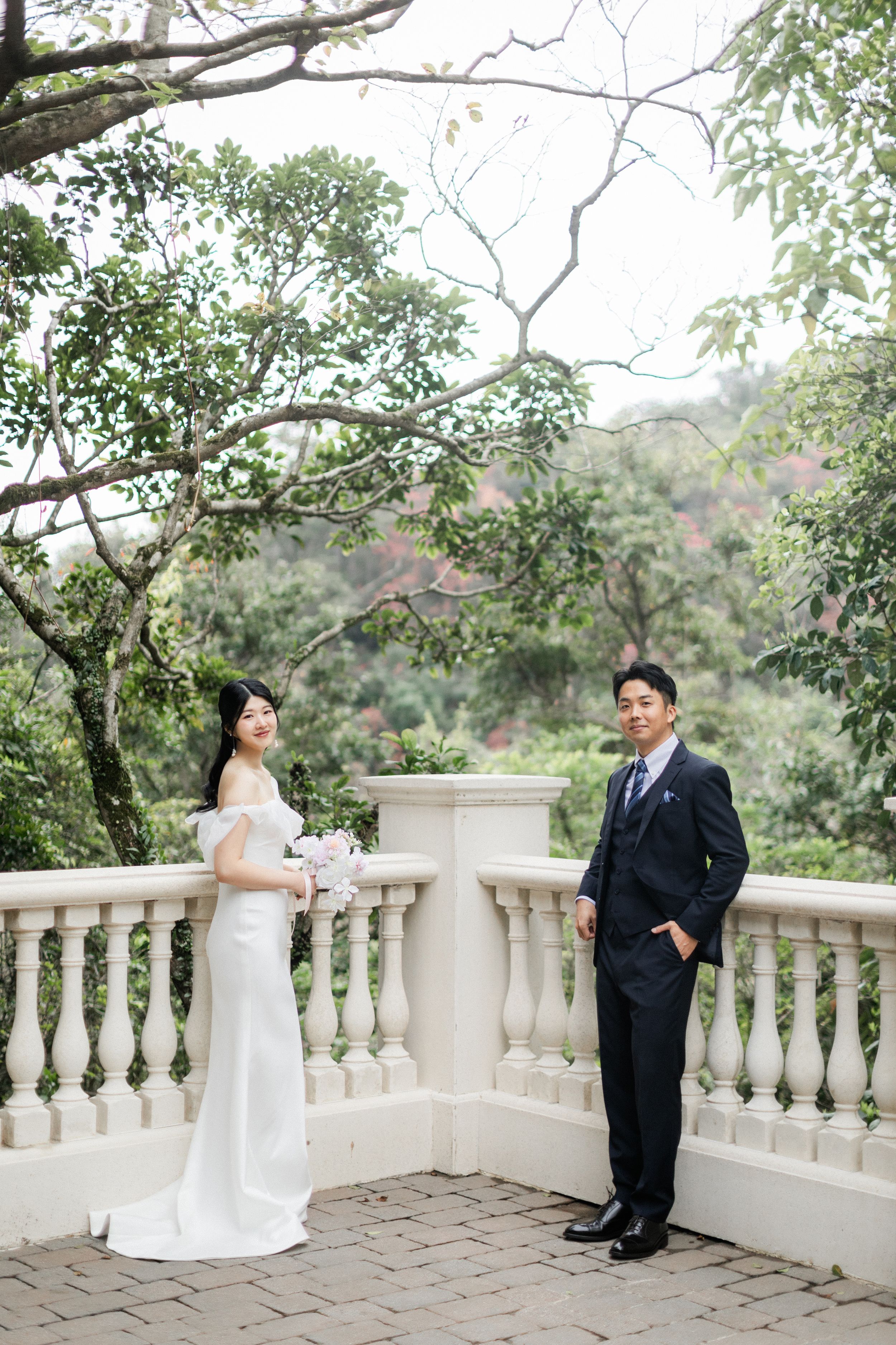 Bride and groom posing apart on a garden terrace, bride in white gown holding bouquet, lush greenery behind.