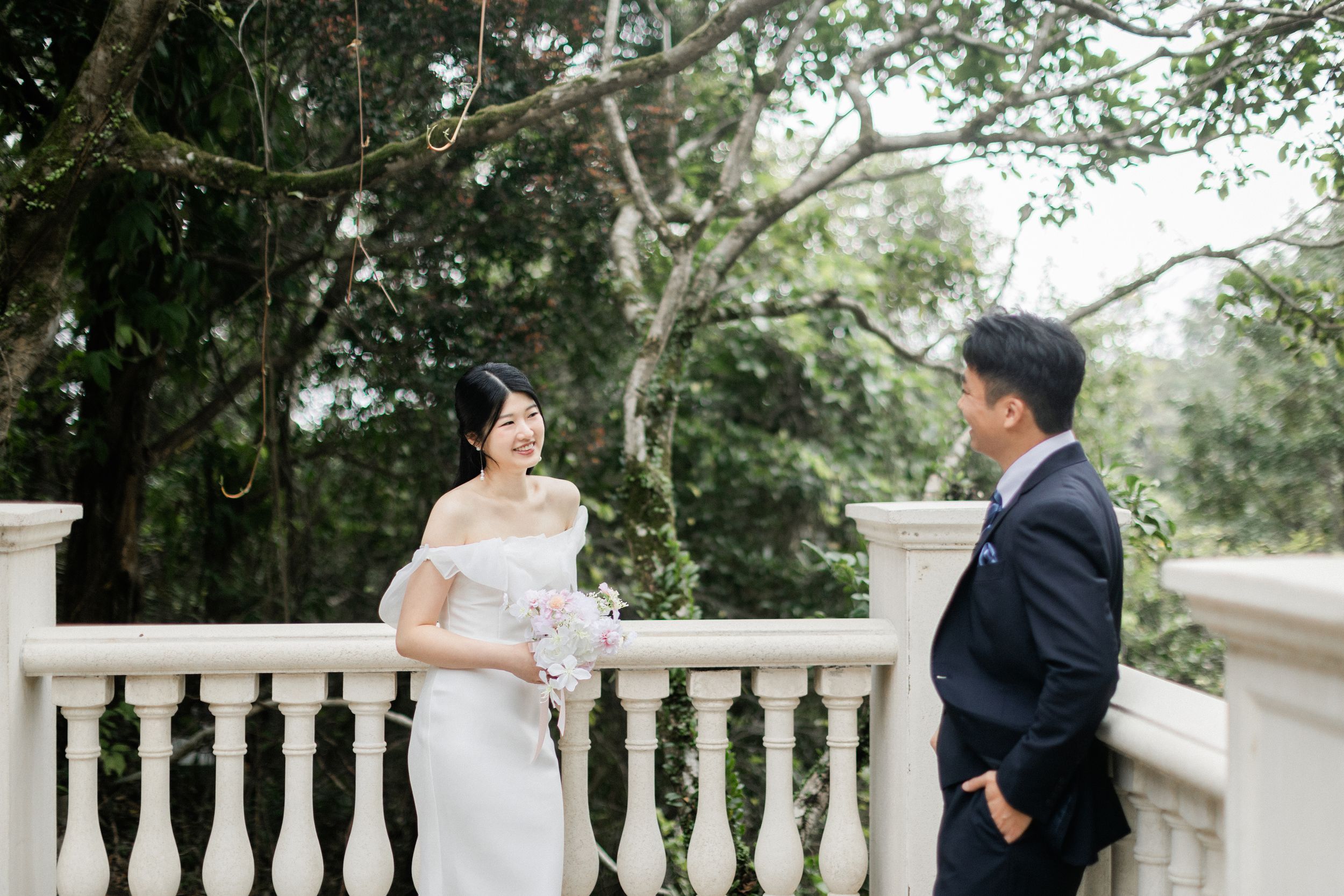 Bride in white off-shoulder gown and groom in navy suit share a romantic moment on a white balustrade terrace.