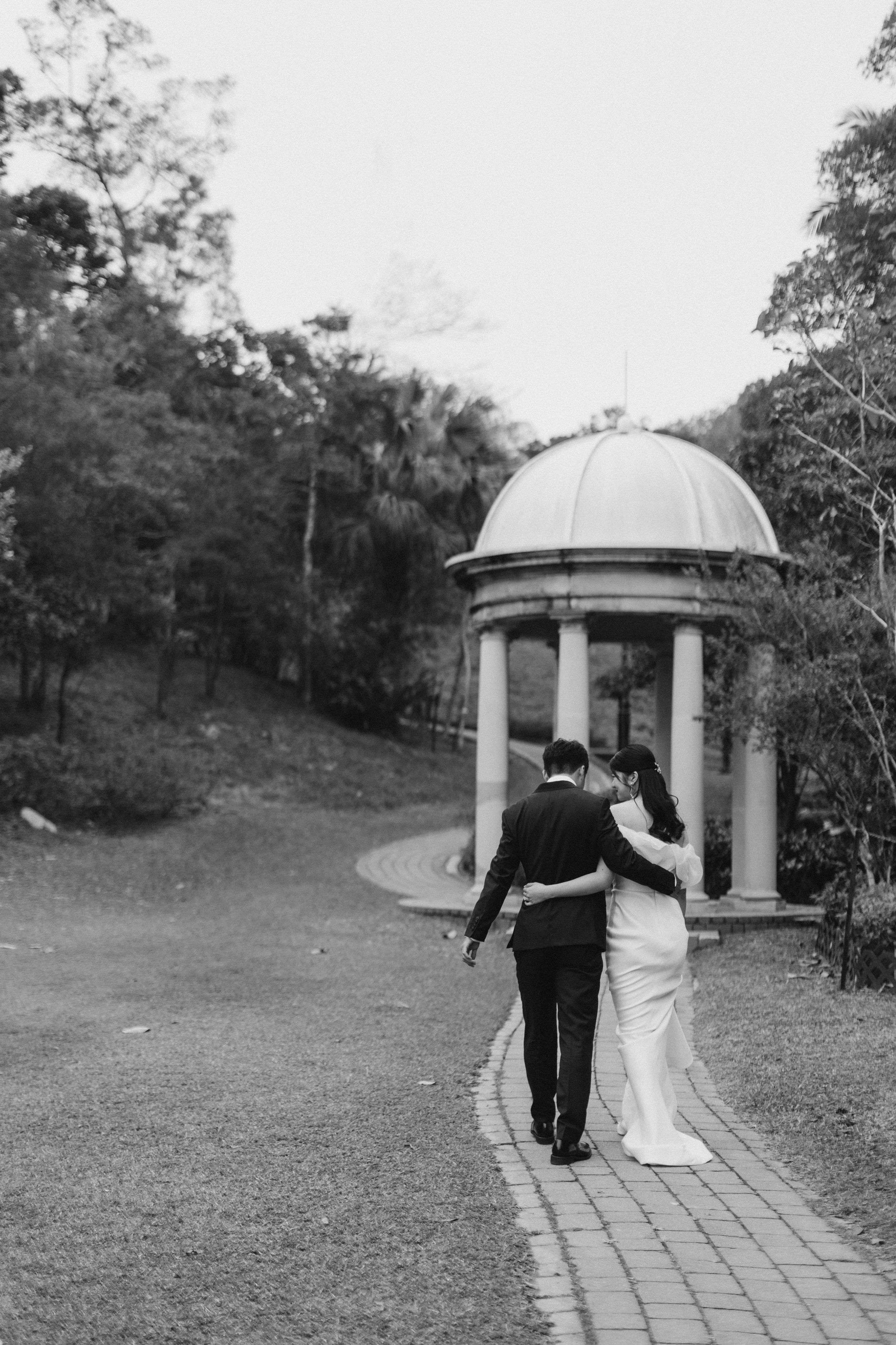Black and white photo of newlyweds walking arm in arm toward a classical domed gazebo in a lush garden.