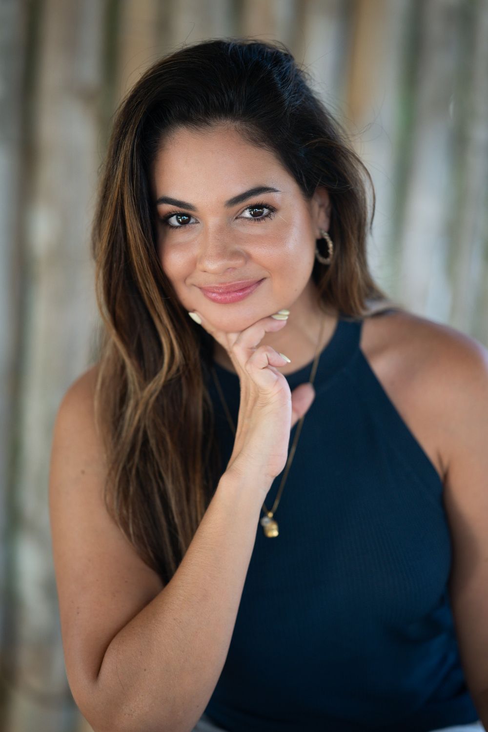Professional portrait of woman in navy sleeveless top with long brown hair posing with hand under chin against blurred background.