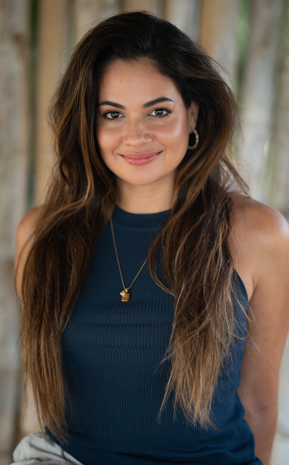 Professional portrait of a person with long brown hair wearing a navy blue sleeveless top and delicate gold necklace.