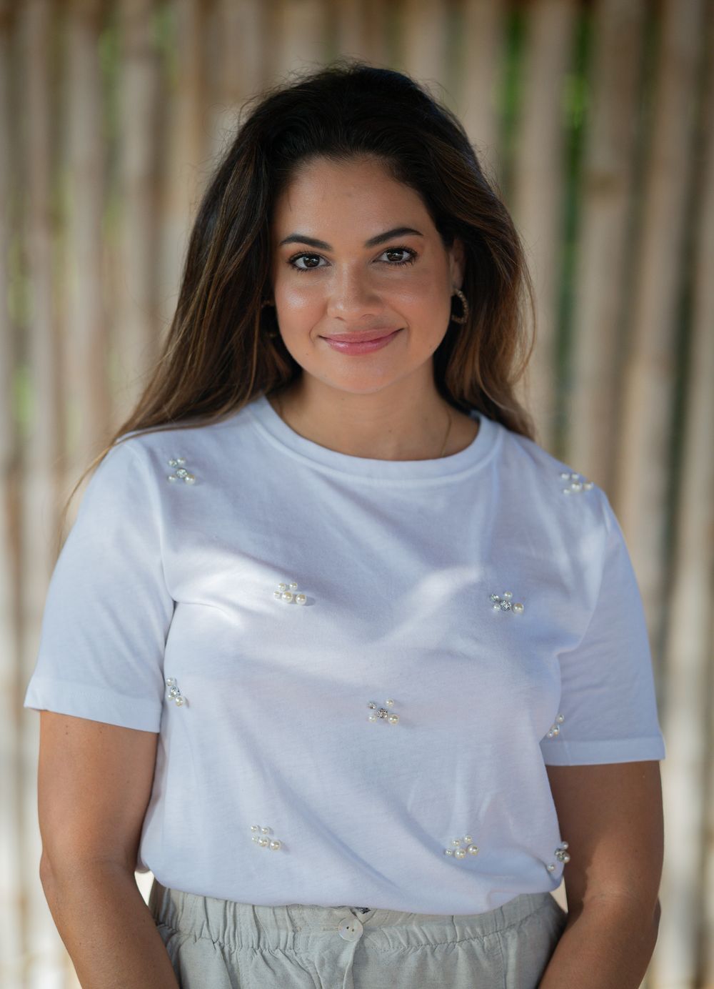 Smiling woman in white t-shirt decorated with small embellishments against bamboo backdrop.