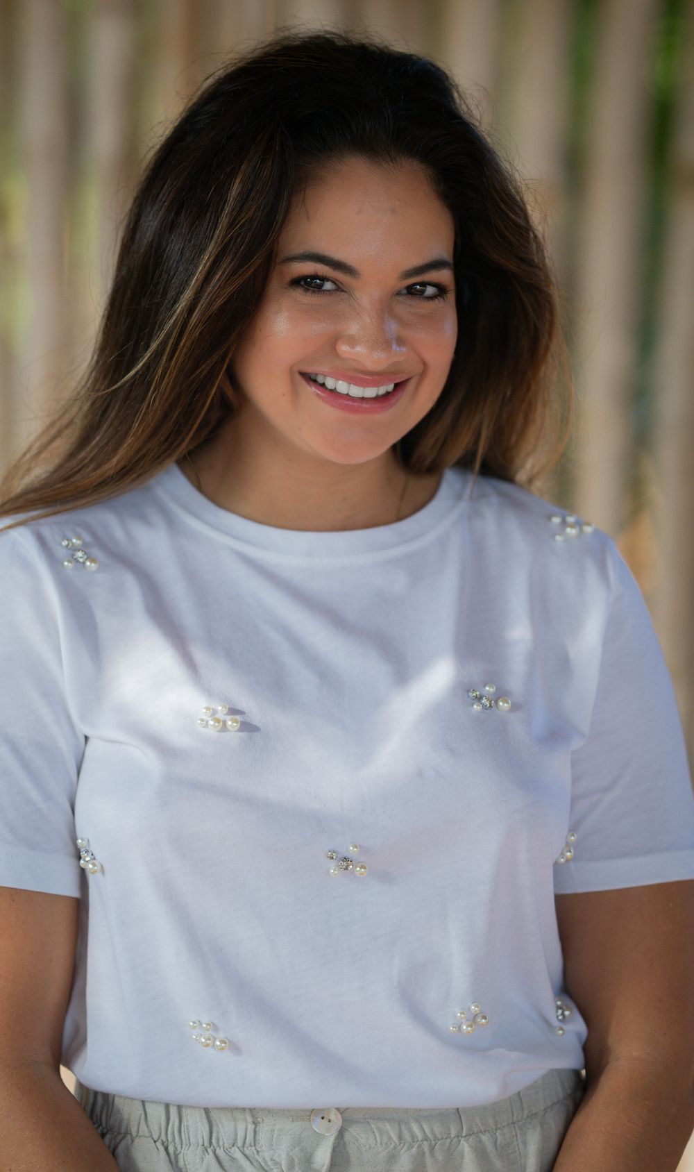 Portrait shot of a smiling woman wearing a white t-shirt with delicate jeweled embellishments against a soft blurred background.