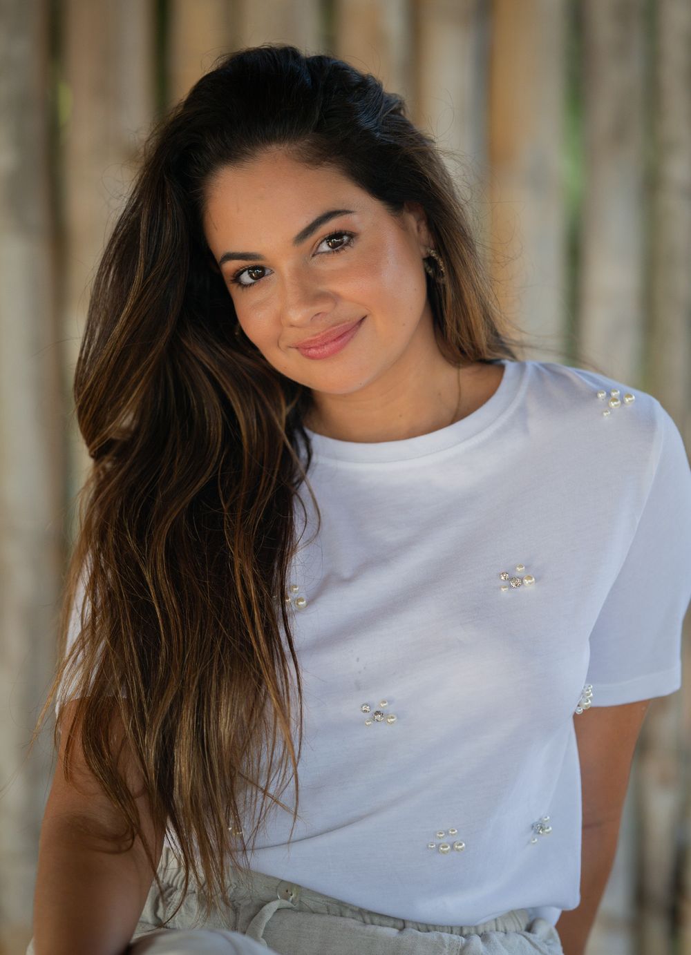 Smiling young model in white t-shirt with decorative details and long brown hair posing against blurred background.