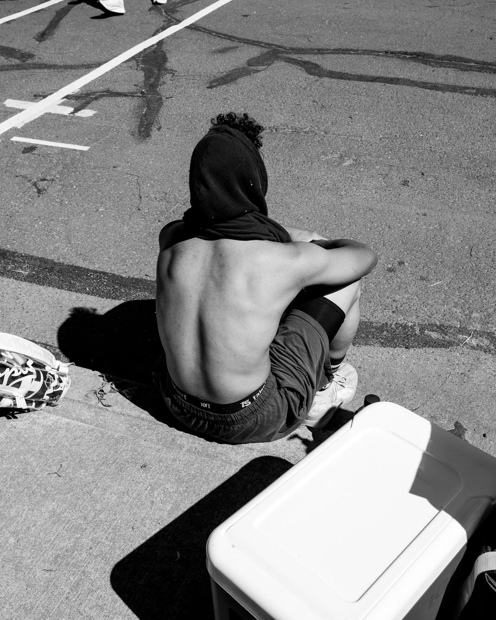 A shirtless person sits on pavement near white lines in black and white photography.