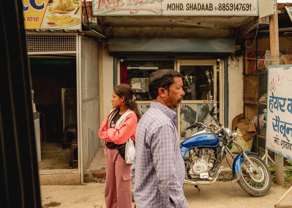 Street scene in India with pedestrians walking past shops and a parked motorcycle.