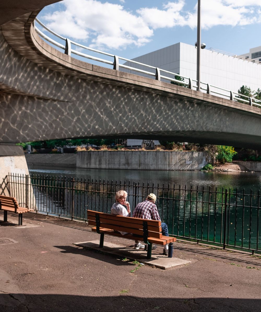 Two people sitting on bench overlooking water under curved bridge on sunny day.