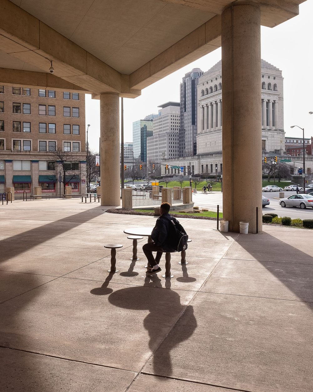 Person sitting alone on bench under concrete columns with cityscape view.
