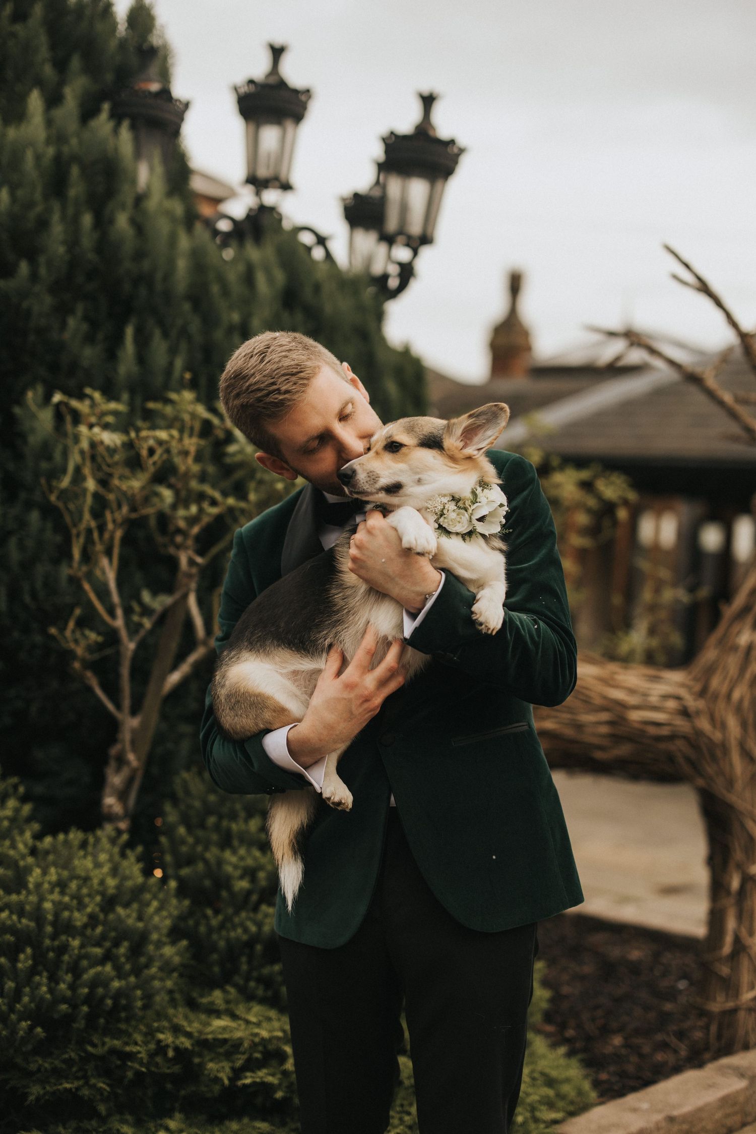 A sweet moment in formal wear with a Corgi dog being embraced outdoors near vintage-style lampposts and greenery.