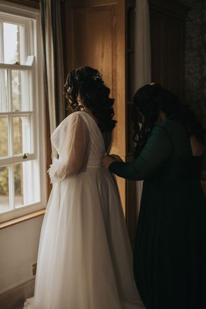 A bride in a flowing white wedding dress stands silhouetted against a sunlit window while getting ready for her ceremony.
