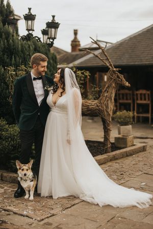 A newlywed couple stands in formal attire on stone paving with a sweet dog and rustic decor in an outdoor garden setting.