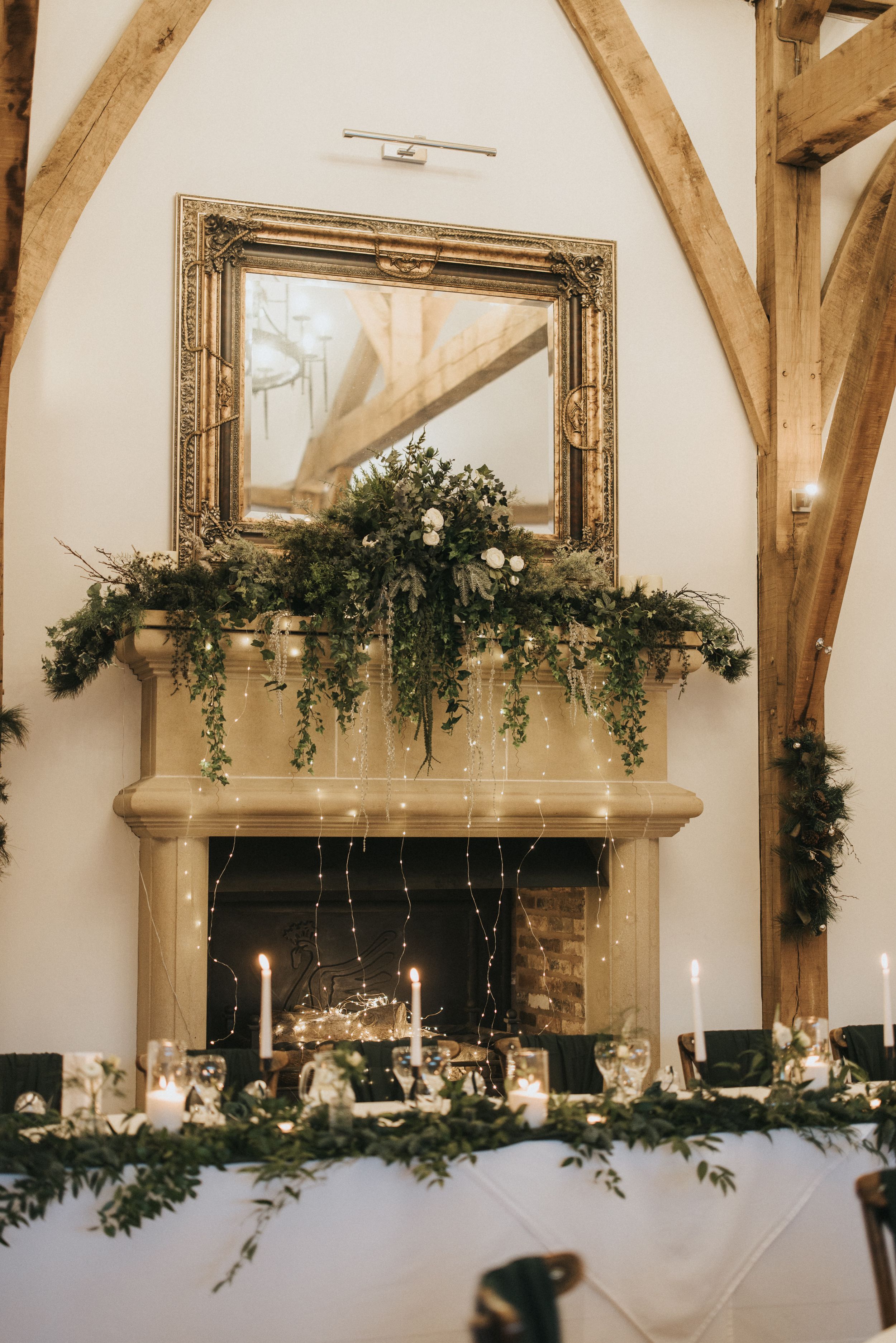 Rustic wooden beams and elegant floral garlands adorn a white wedding reception venue with vintage mirrors and candles.