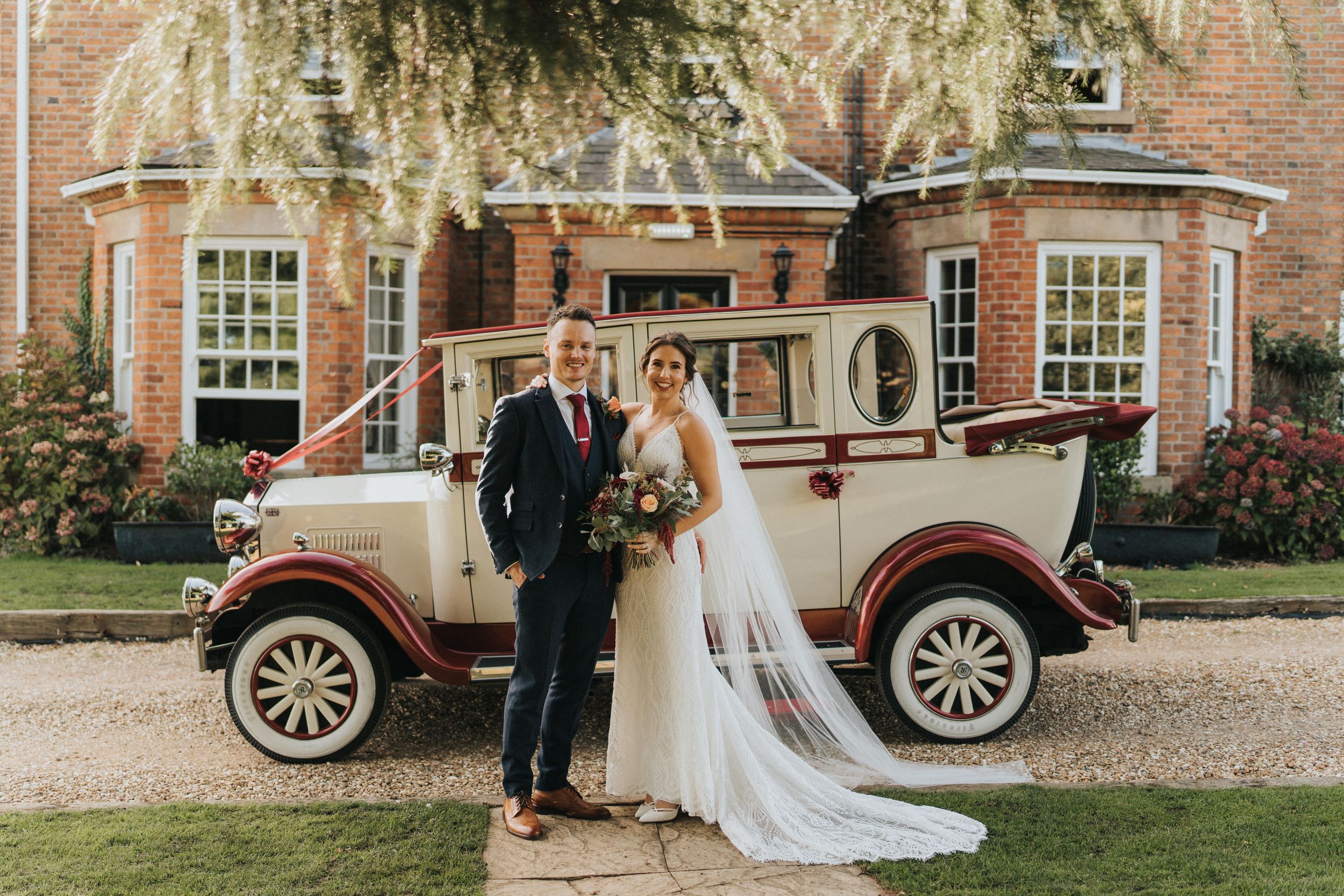 Couple in formal wedding attire stands next to vintage car at brick building during golden hour photography session.