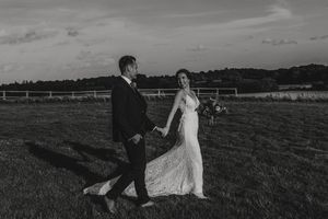 A wedding couple walks hand in hand across a grassy countryside field during sunset in black and white photography.